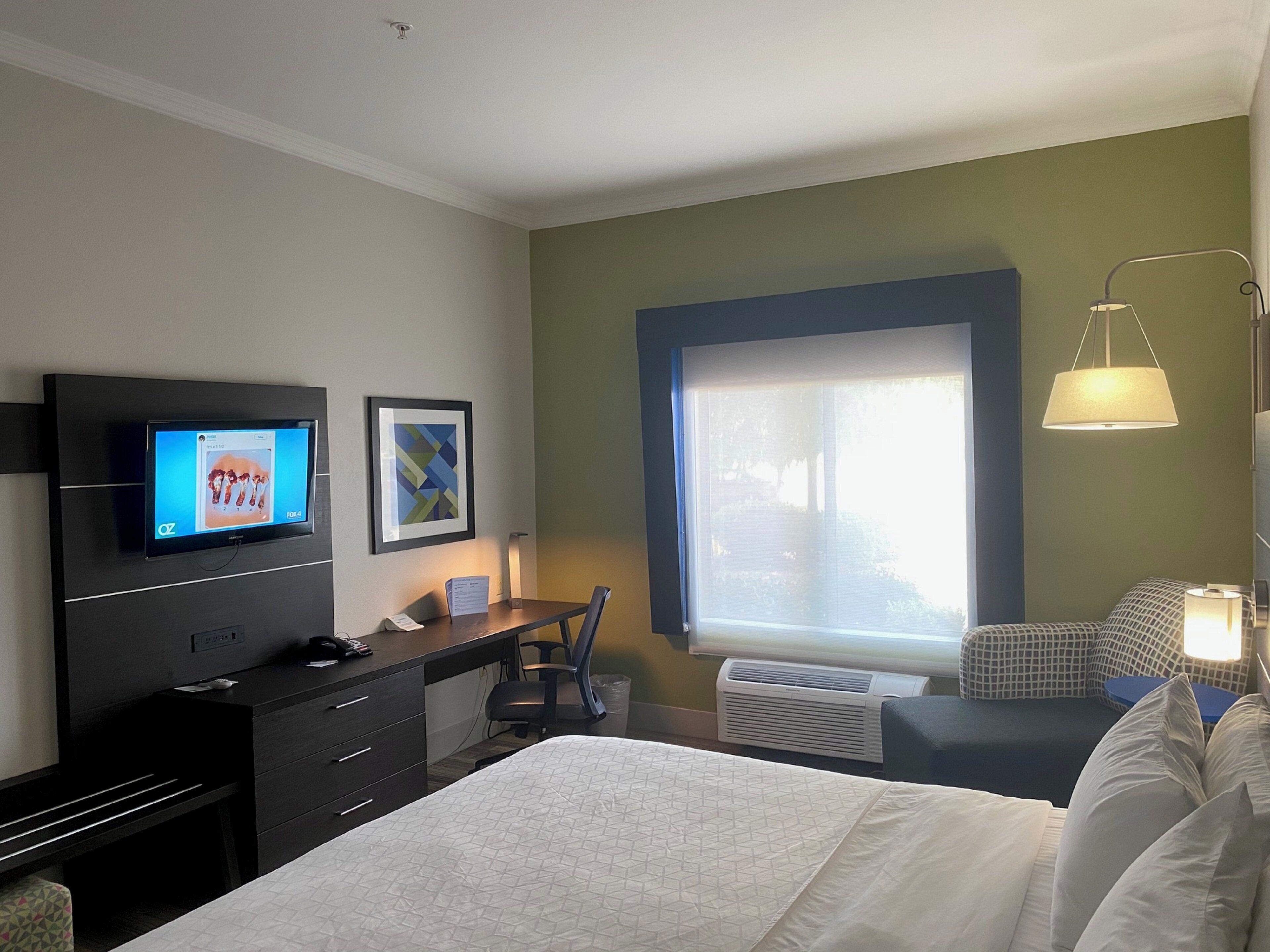 A hotel room with a bed, desk, chair, TV, artwork, window, and lamp.