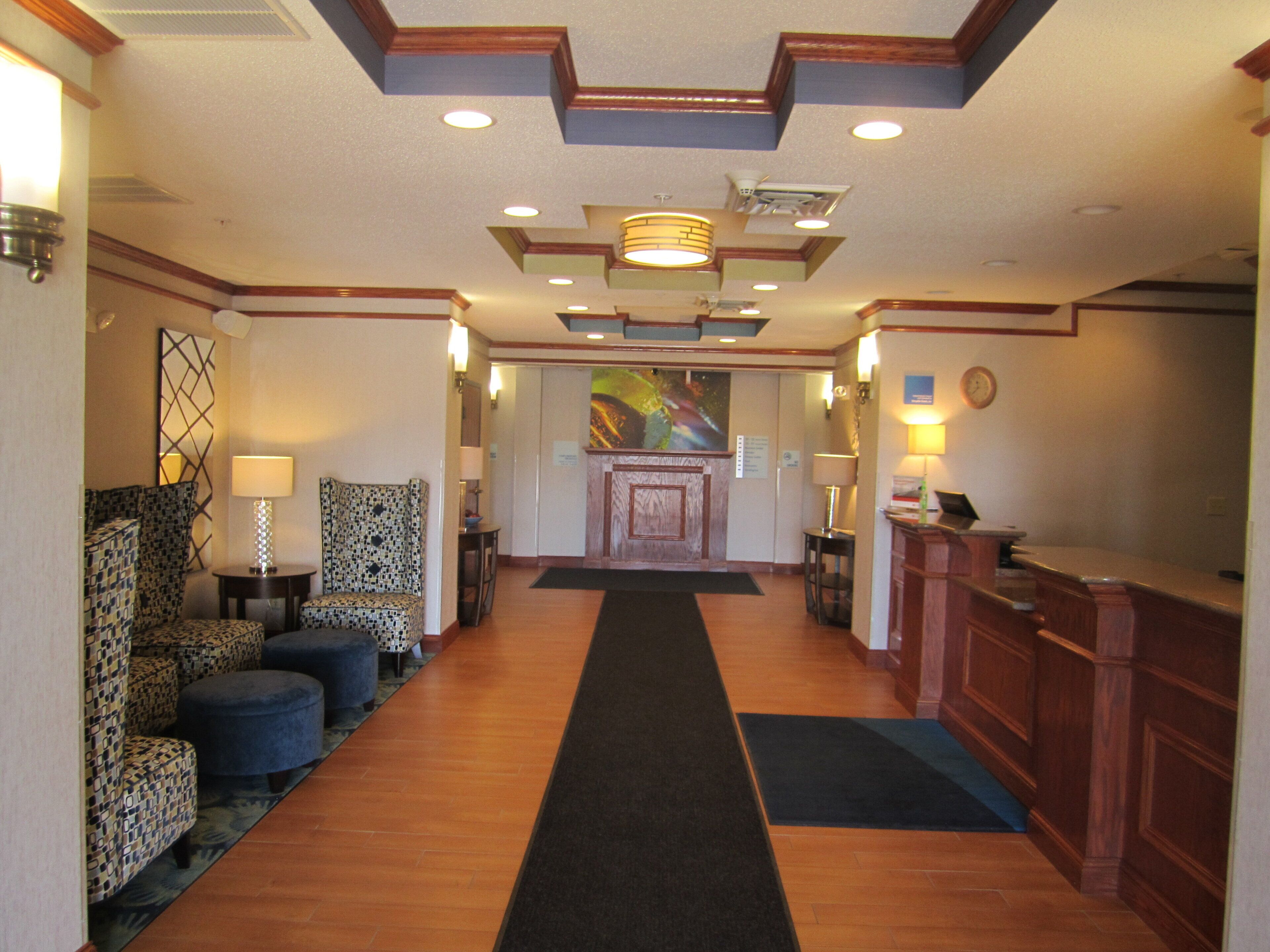 A hotel lobby with a reception desk, patterned chairs, a long carpeted walkway, and a wall-mounted clock.
