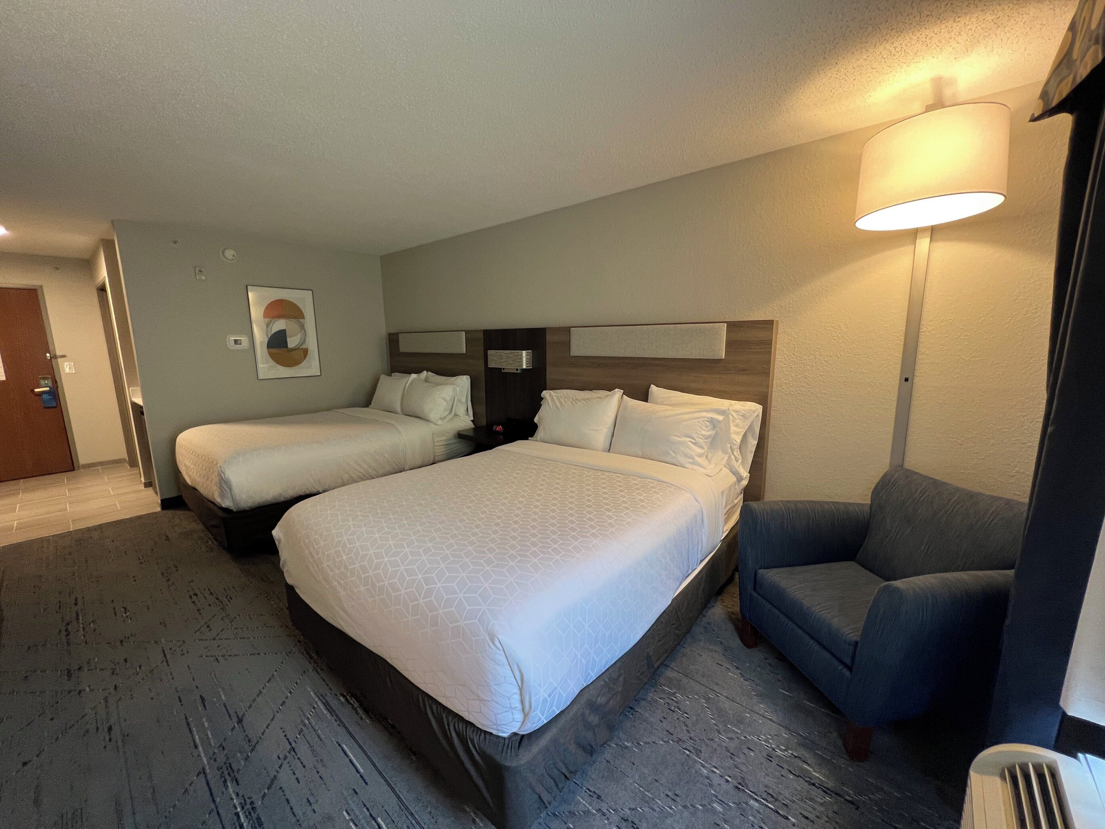 Quarto de hotel com duas camas, um sofá, uma luminária fixada na parede e uma porta.