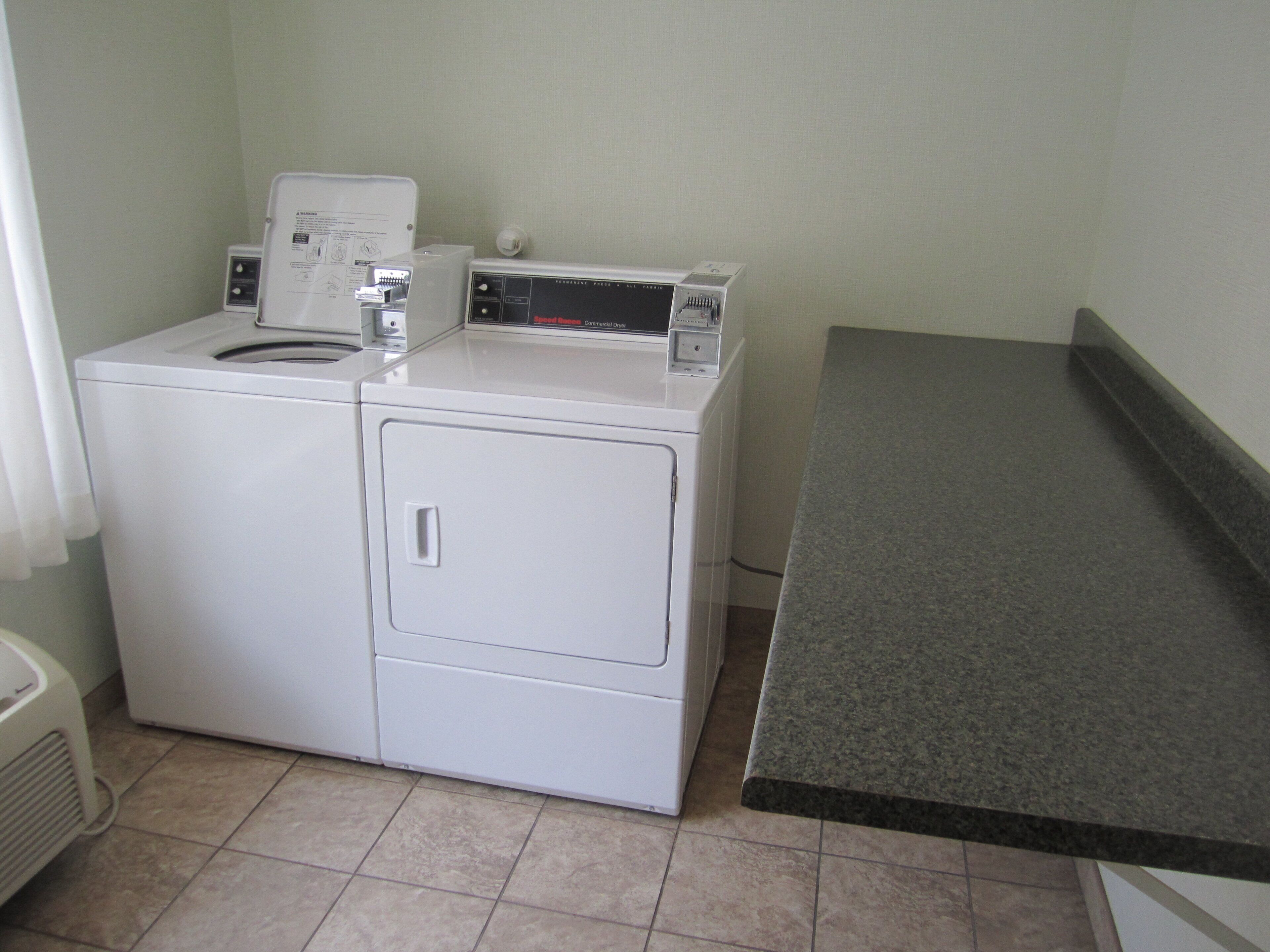 A compact, white, front-loading washer and dryer unit with a control panel on top, situated in a corner of a room with a grey countertop and tiled floor.