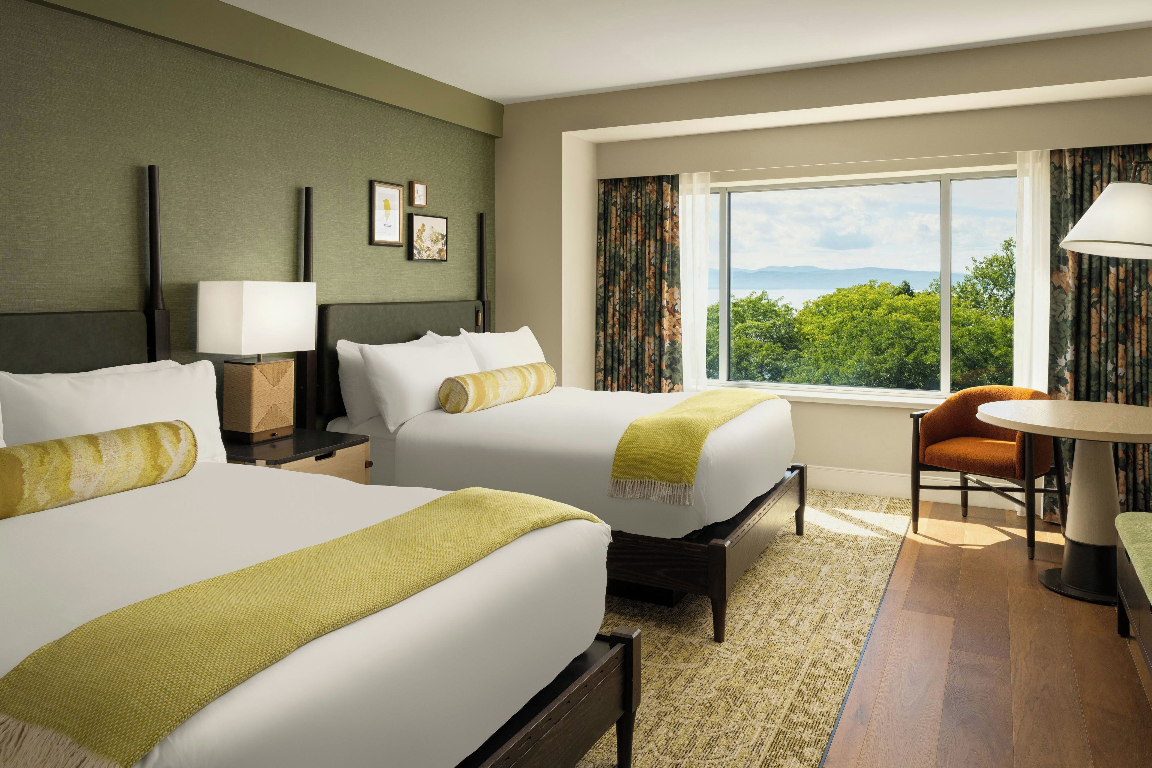 A hotel room with two beds, a desk, a chair, and a view of the outdoors.