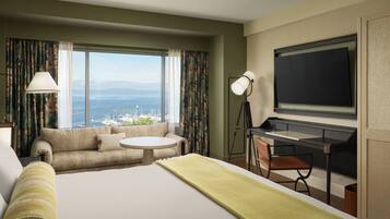 A hotel room with a large bed, a desk, a chair, a round table, a television, and a view of the sea.