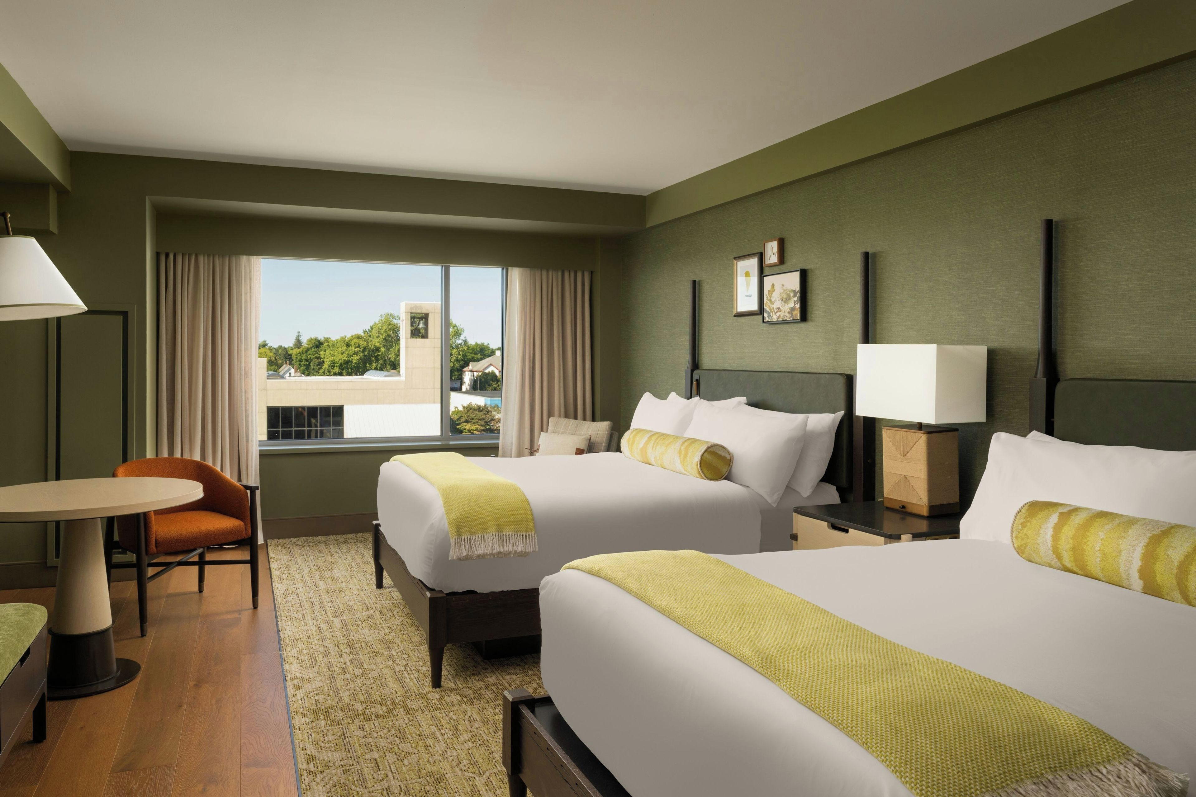 A hotel room with two beds, a desk, a chair, a window with curtains, and a view of the outside.