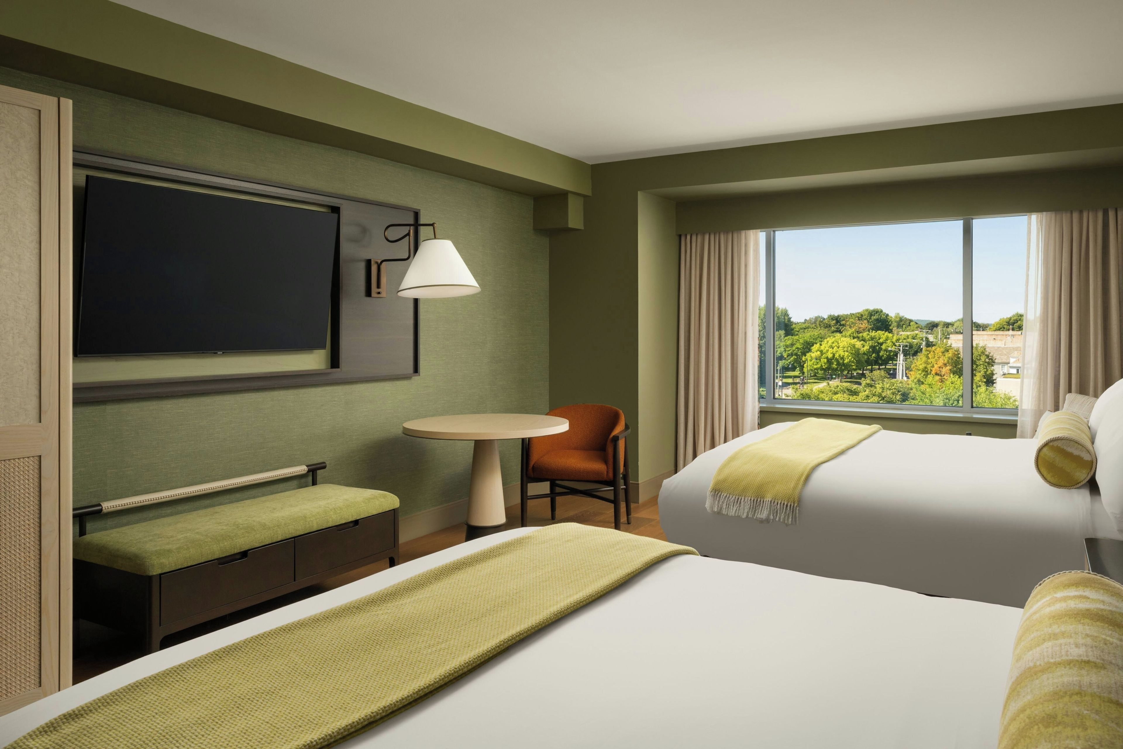 A hotel room with a bed, a desk, a chair, a TV, and a window with a view of trees.