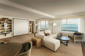 A modern living room with a sofa, armchairs, a coffee table, a bookshelf, and a large window with a view of the sea. - The Harborvale, Autograph Collection (Burlington)