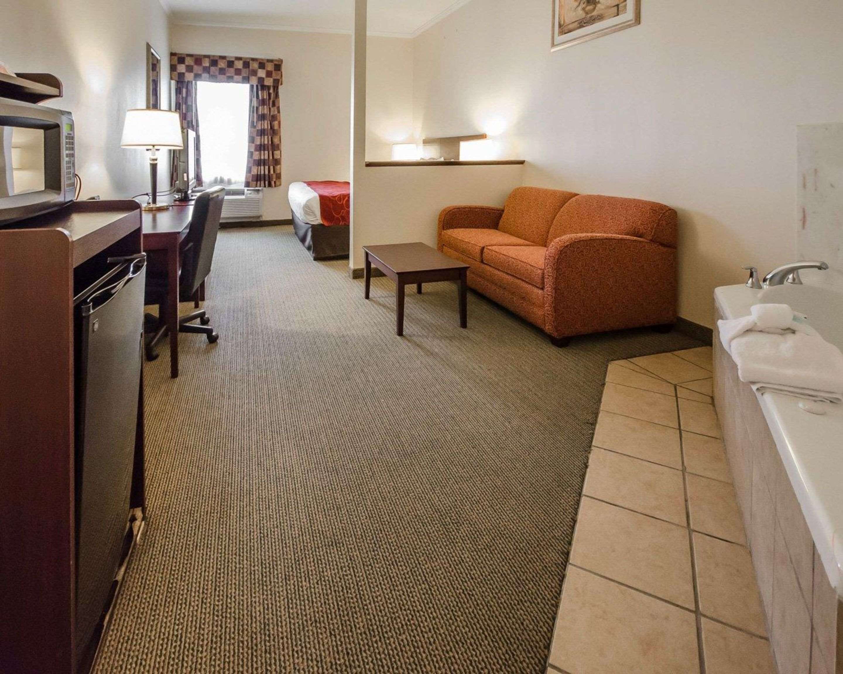 A hotel room with a kitchenette, a sofa, a small table, a lamp, and a bathroom with a sink and mirror.