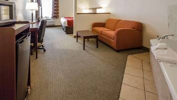 A hotel room with a kitchenette, a sofa, a small table, a lamp, and a bathroom with a sink and mirror.