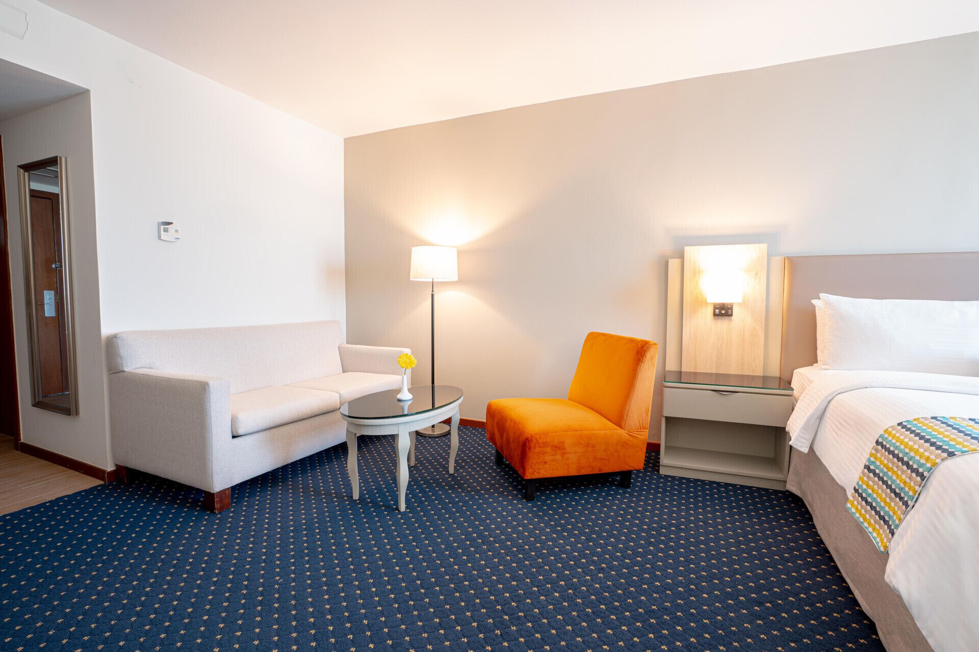 Quarto de hotel com sofá, poltrona, mesa pequena, luminária, espelho e cama.