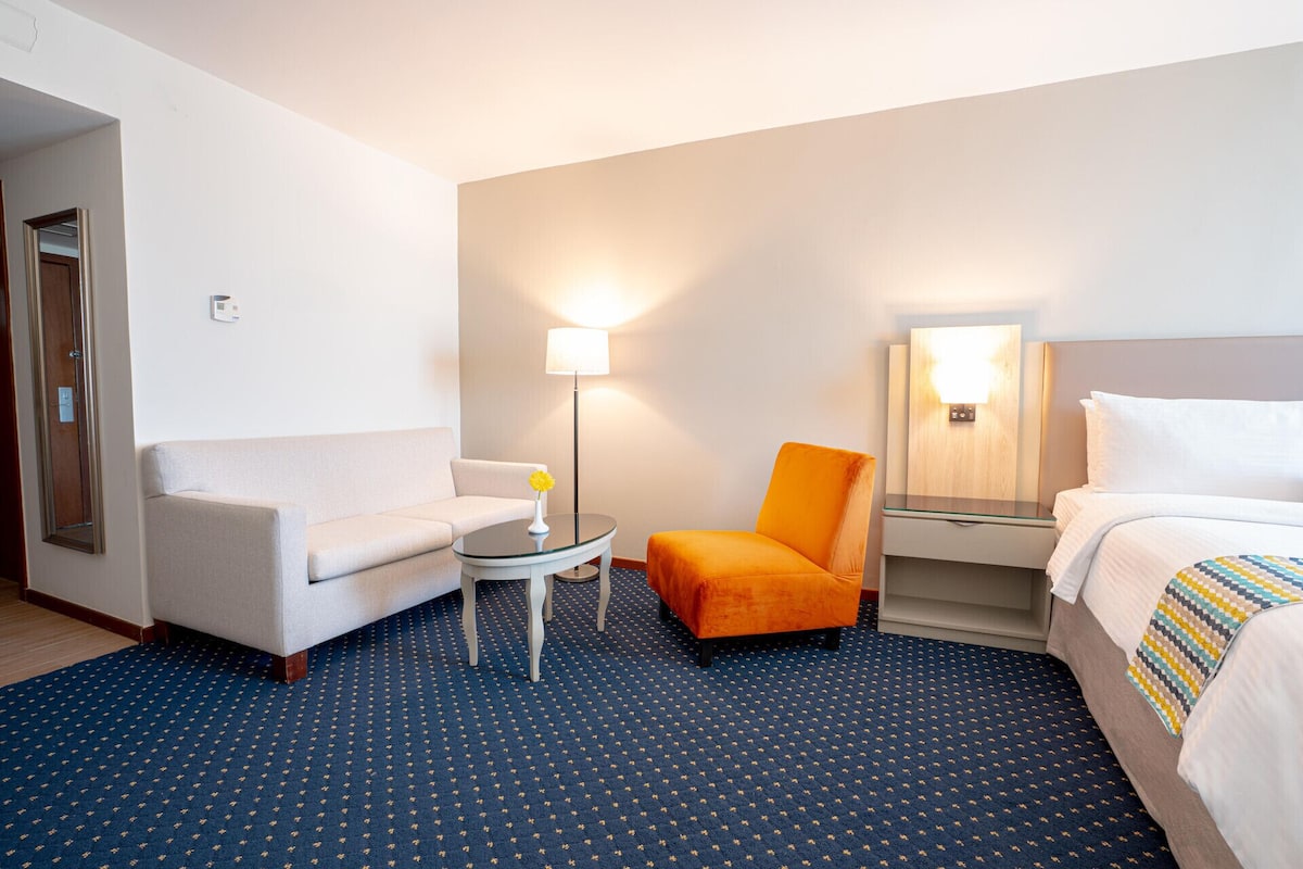 Habitación de hotel con sofá, sillón, una mesita, lámpara, espejo y cama.