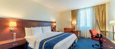 A hotel room with a large bed, two bedside tables, a desk, a chair, and a window with curtains.