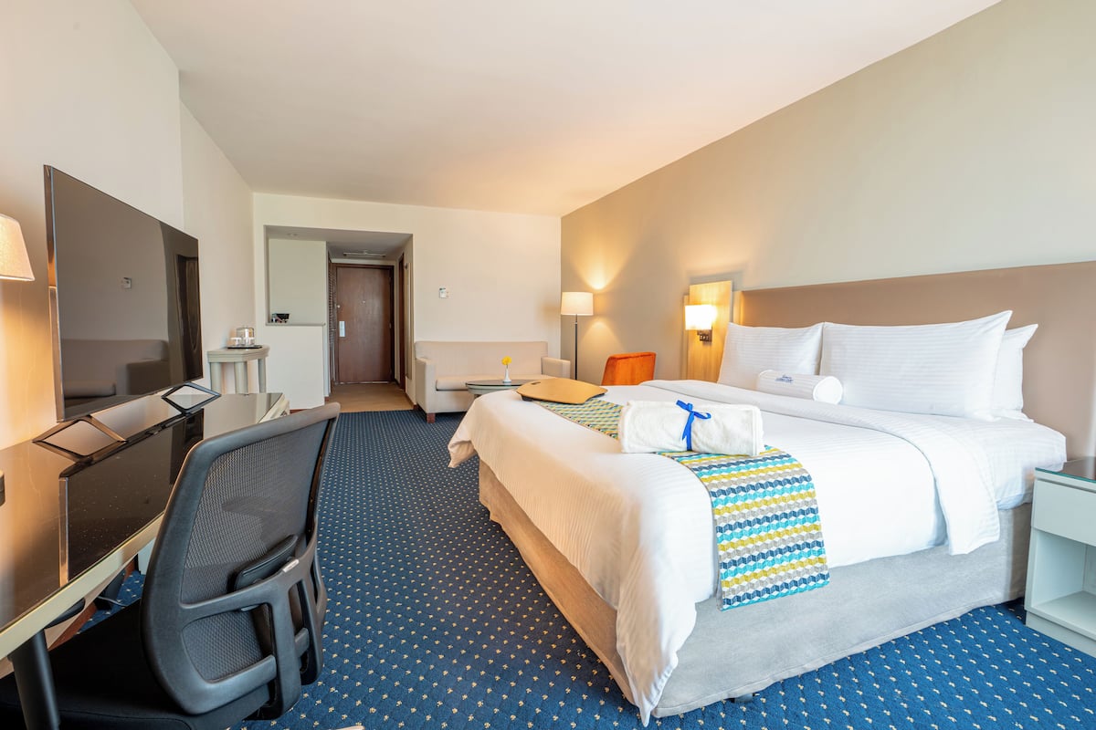 Quarto de hotel com cama, escrivaninha, cadeira e pequena mesa com abajur.