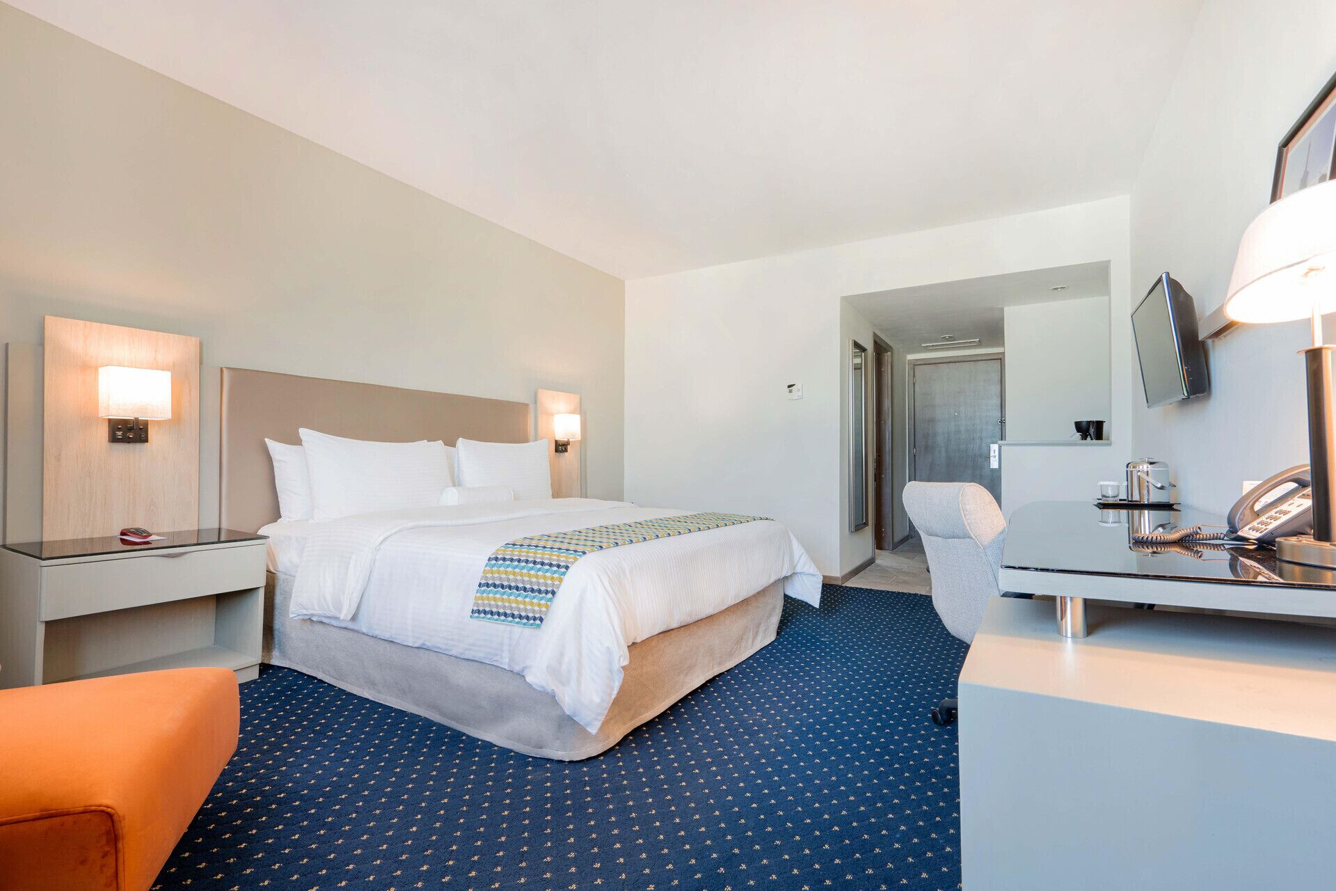 Quarto de hotel com cama, mesa de cabeceira, cadeira, escrivaninha e espelho.