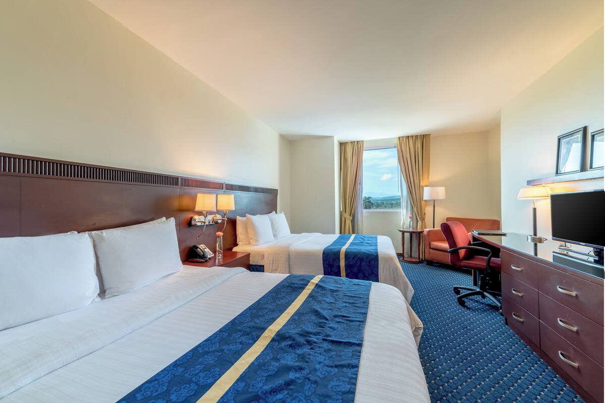 Quarto de hotel com duas camas, uma escrivaninha, uma cadeira, uma televisão e uma janela com cortinas.