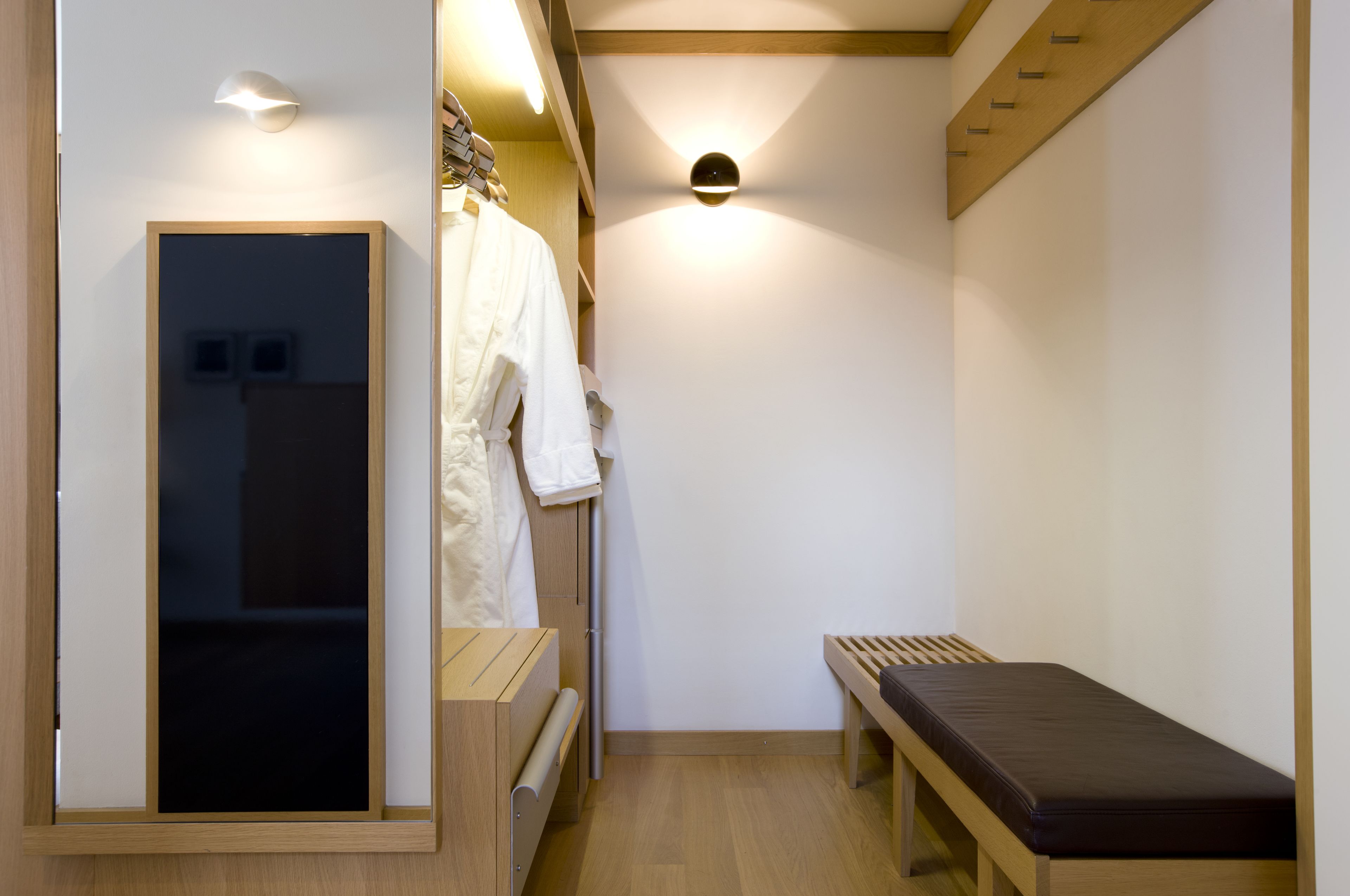 A narrow hallway with a wooden bench, a coat rack with a white robe, and a wall-mounted light fixture.