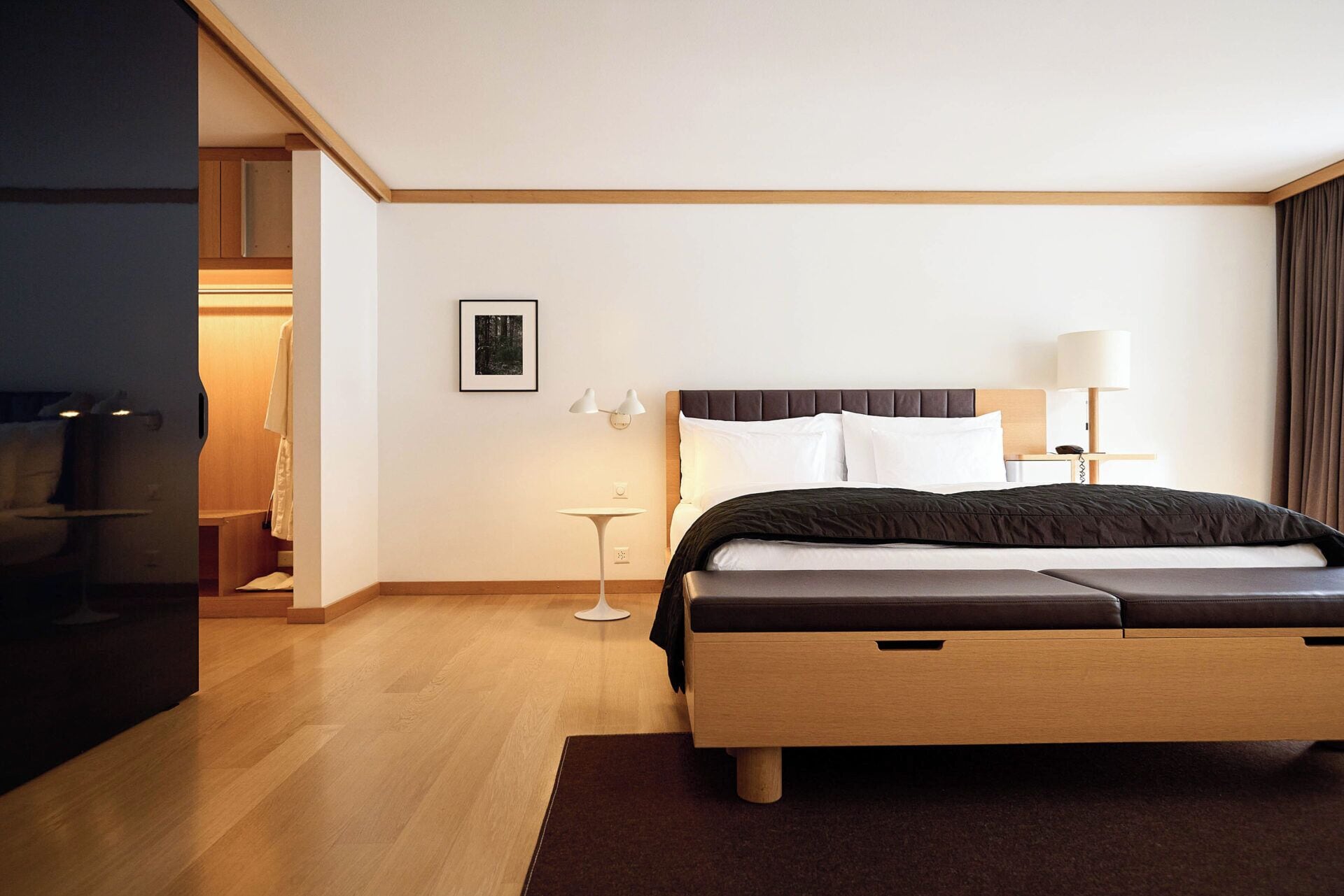 A modern hotel room with a wooden bed, a bedside table with a lamp, a small side table, and a framed picture on the wall.