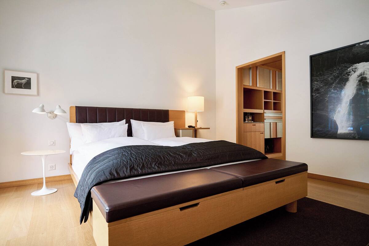 Una habitación de hotel moderna con una cama grande, mesitas de noche, una pequeña mesa redonda, una estantería y un cuadro grande enmarcado en la pared.