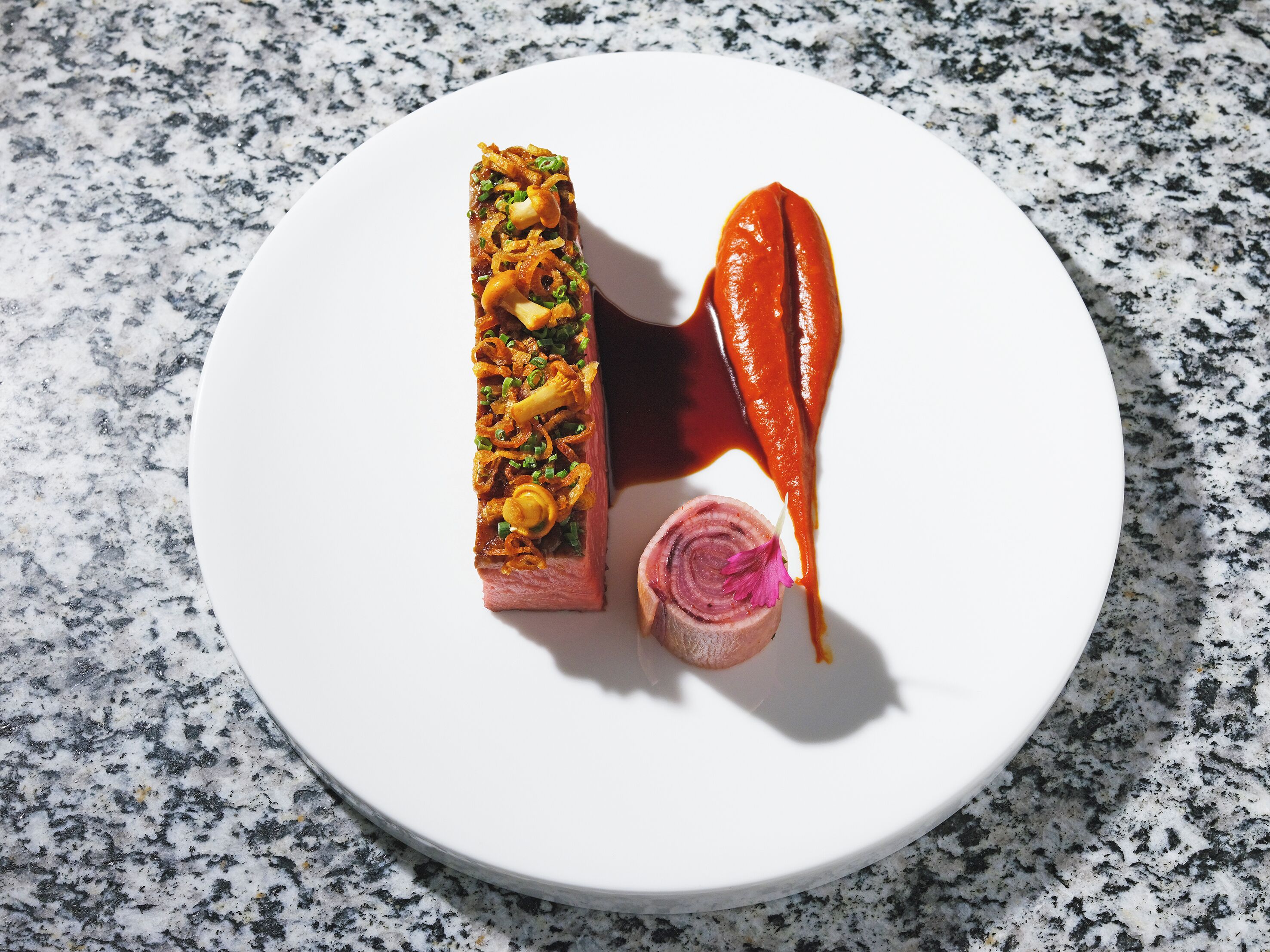 A white plate with a rolled meat dish, a slice of red meat, and a small flower garnish.