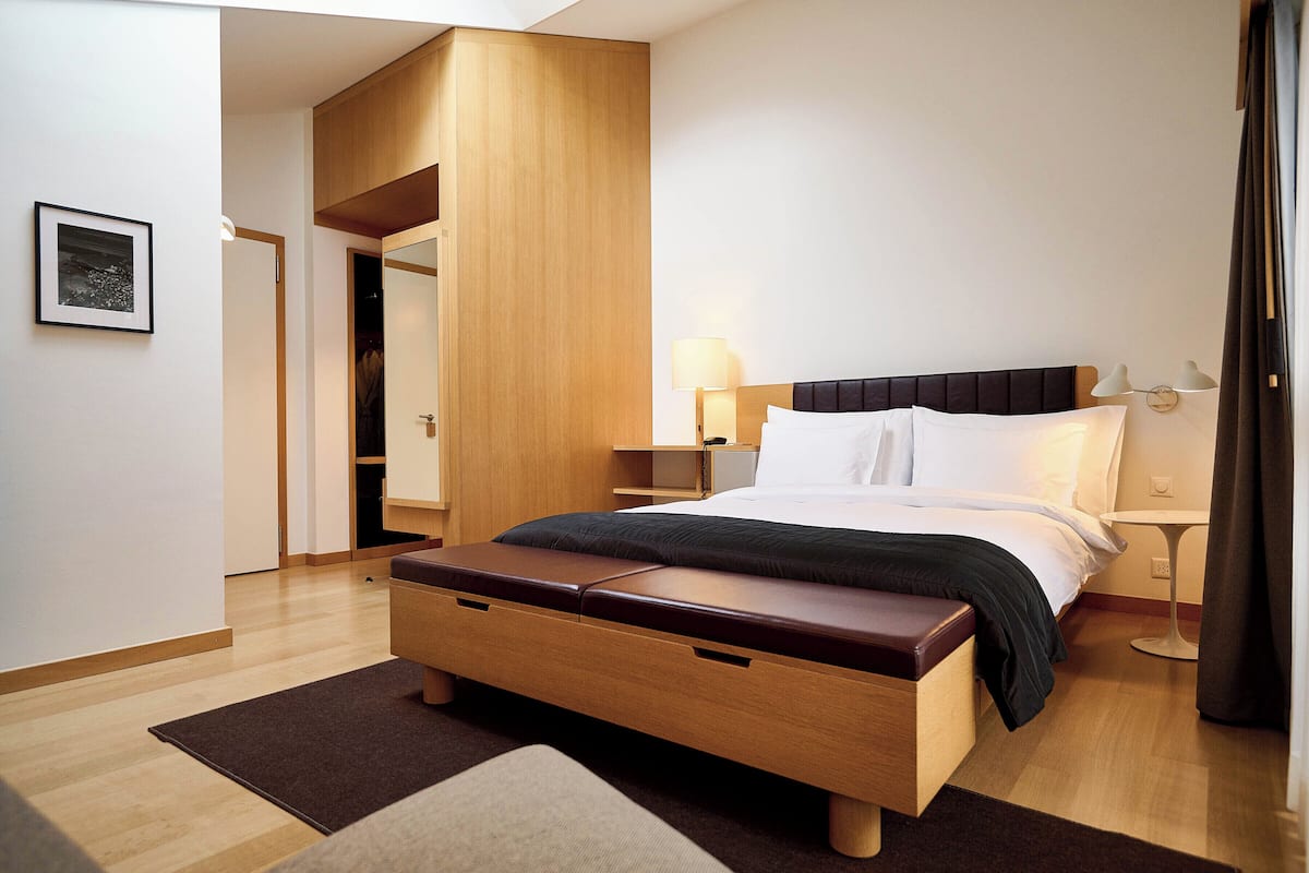 Una habitación de hotel moderna con una cama de madera, una mesita de noche con lámparas y un amplio ventanal con cortinas.