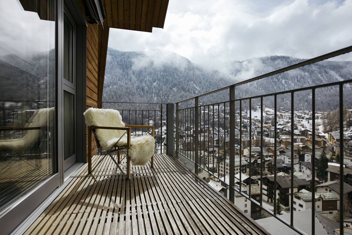 Un balcón con piso de madera y una silla blanca y mullida, con vista a un pueblo nevado y montañas.