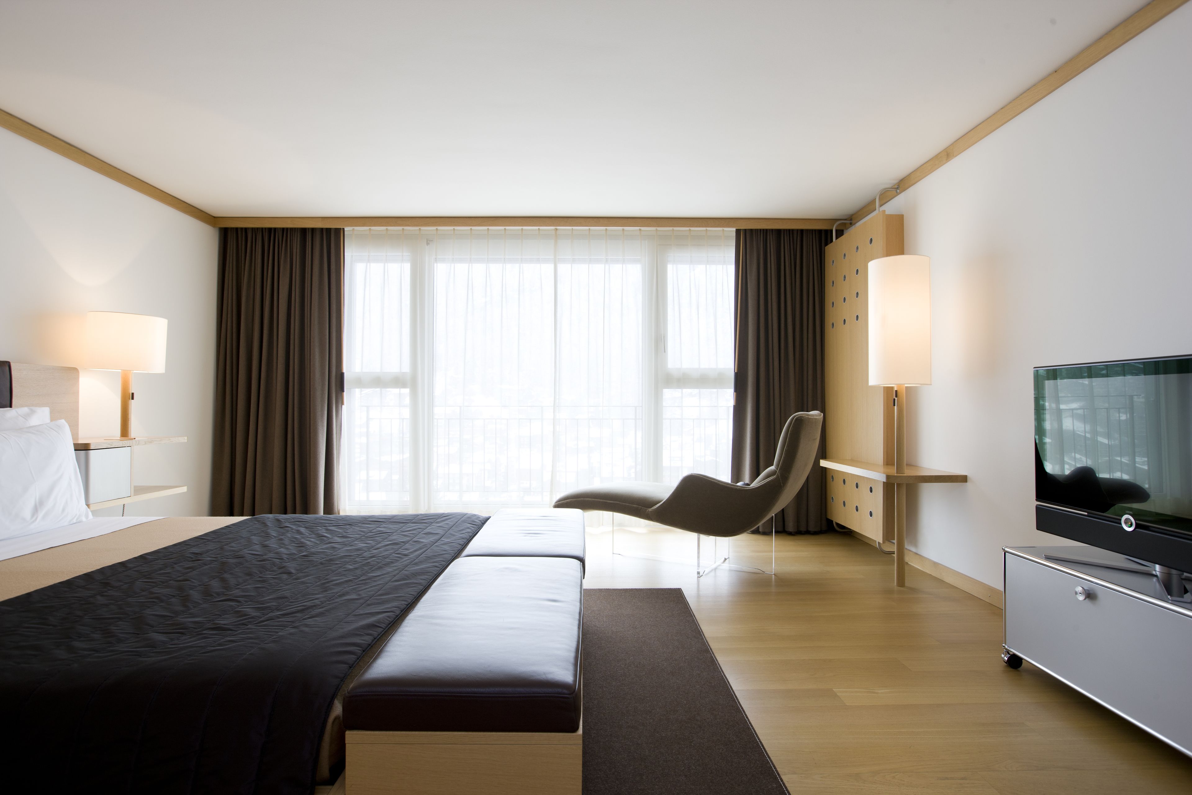 Habitación de hotel con una cama grande, un escritorio con lámpara, una silla, un televisor y un amplio ventanal con cortinas.