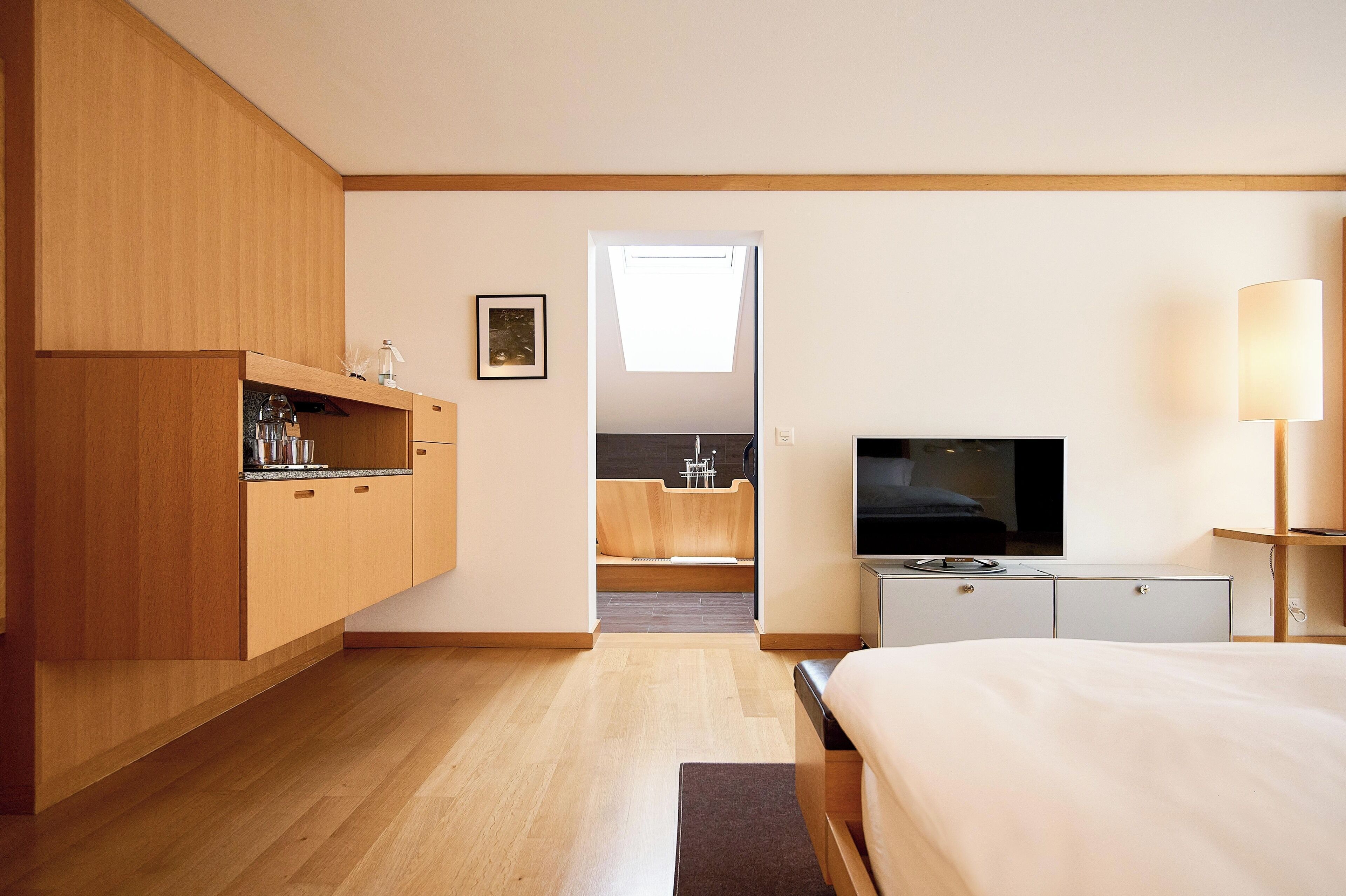 A modern hotel room with a wooden wardrobe, a flat-screen TV, a bedside lamp, and a view of a kitchen and bathroom through an open door.