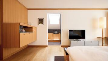A modern hotel room with a wooden wardrobe, a flat-screen TV, a bedside lamp, and a view of a kitchen and bathroom through an open door.