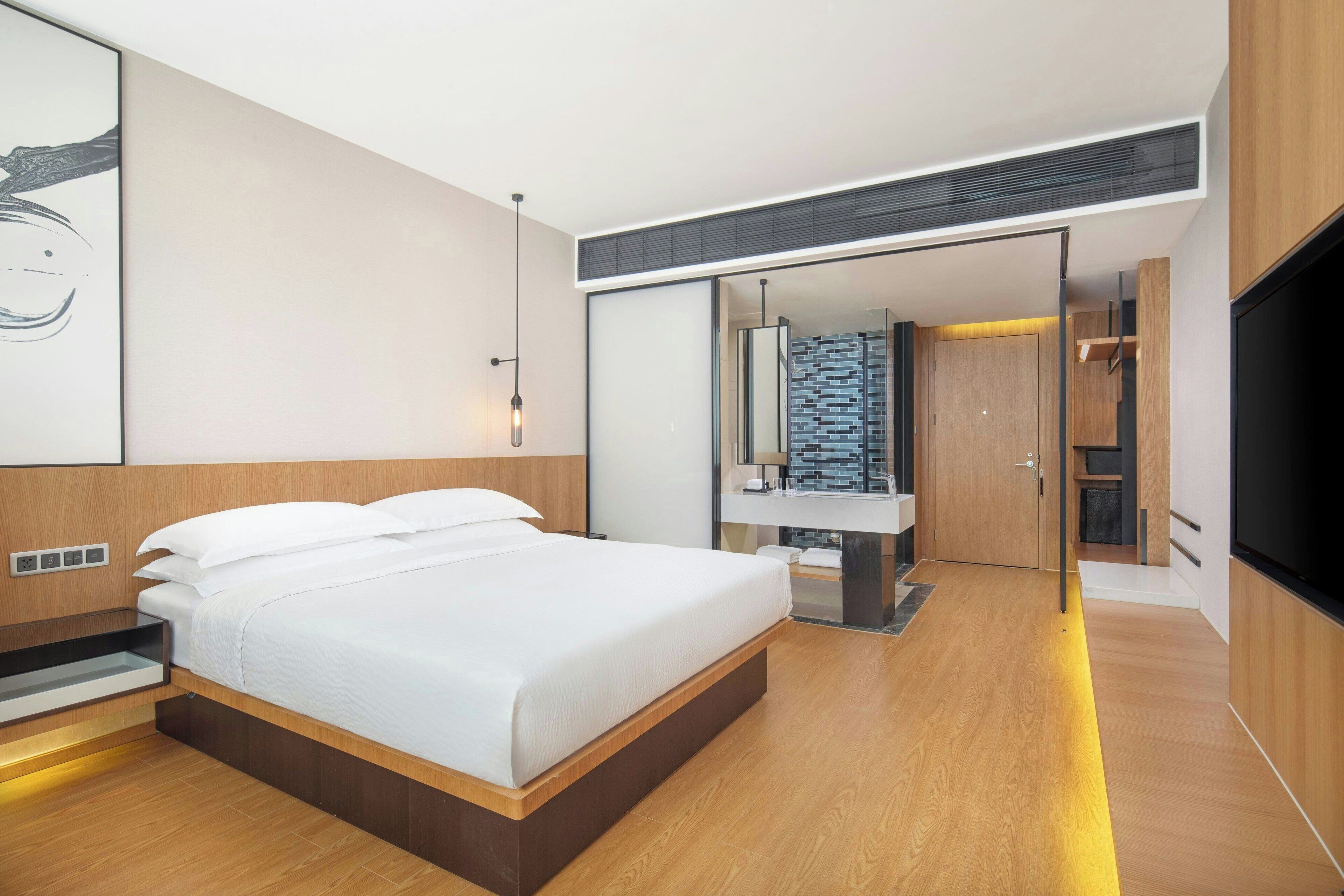A modern bedroom with a large bed, a bedside table, a mirror, and a bathroom visible through an open door.