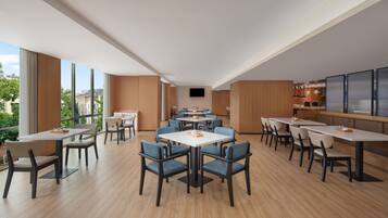 A spacious dining area with multiple tables and chairs, large windows offering a view of the city, and a modern kitchen in the background.