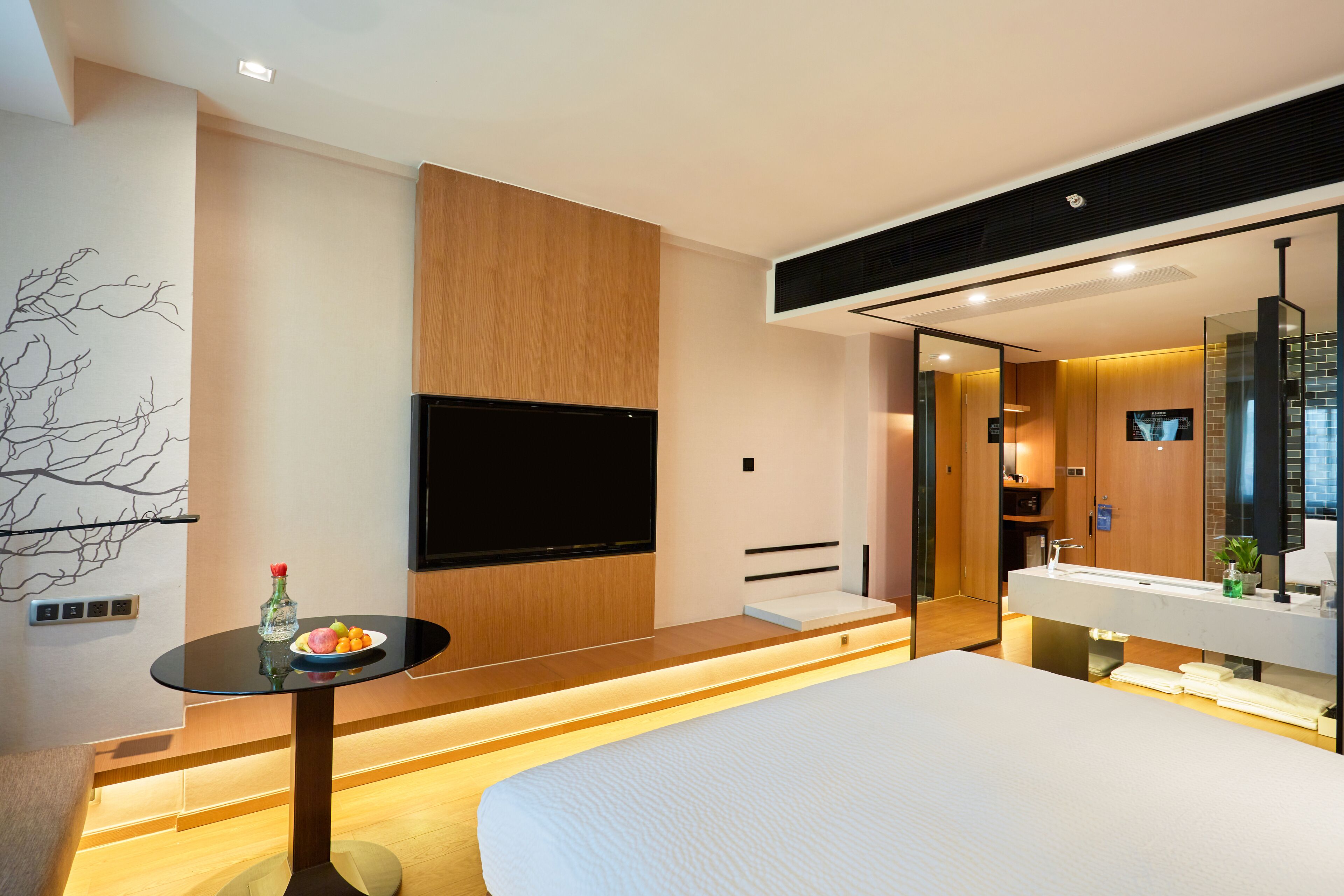Ein modernes Hotelzimmer mit einem großen Bett, einem Flachbildfernseher, einem kleinen Tisch mit Obstschale und einem Badezimmer mit großem Spiegel und Waschbecken.