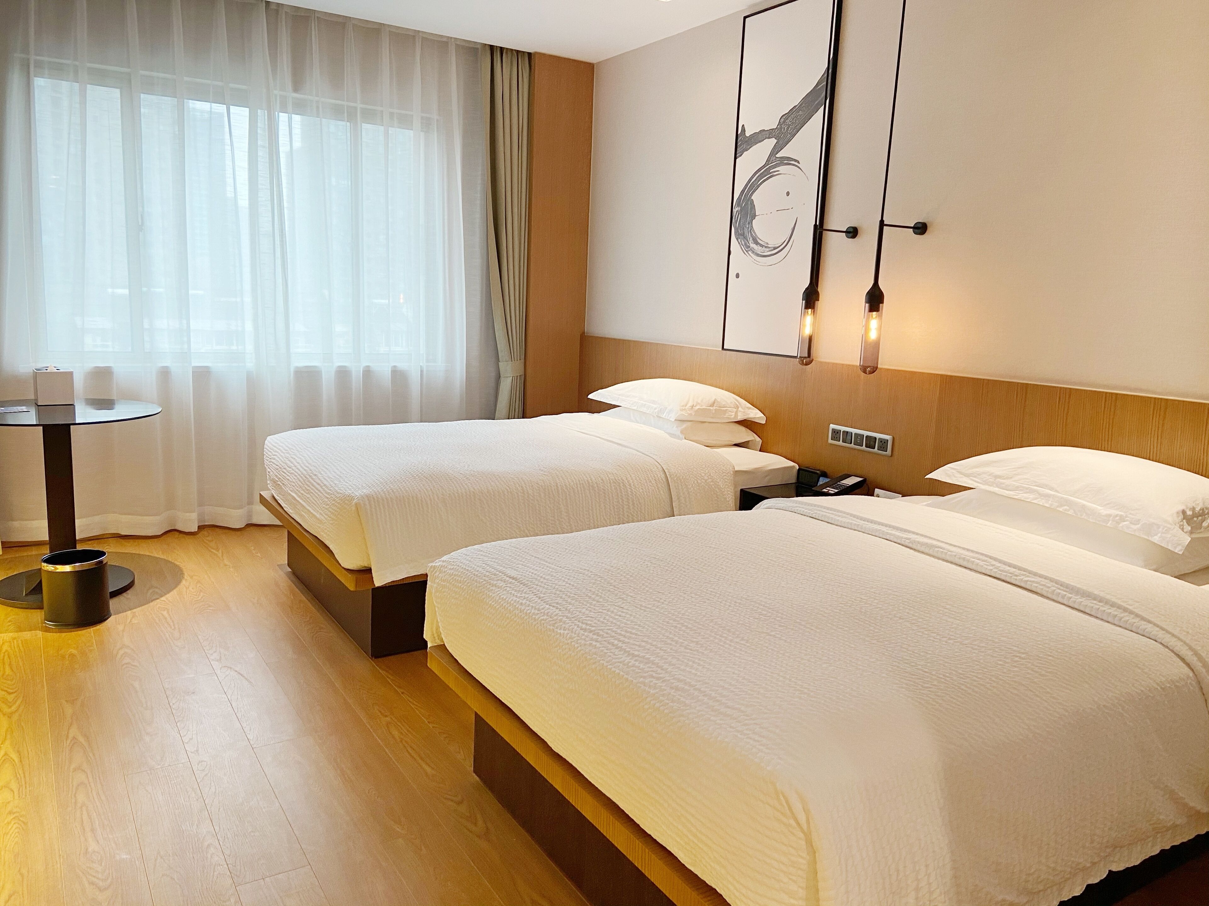 A hotel room with two beds, a small table, a lamp, and a wall-mounted artwork.