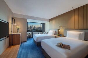 Habitación de hotel con dos camas, un escritorio y un ventanal con vistas a la ciudad.