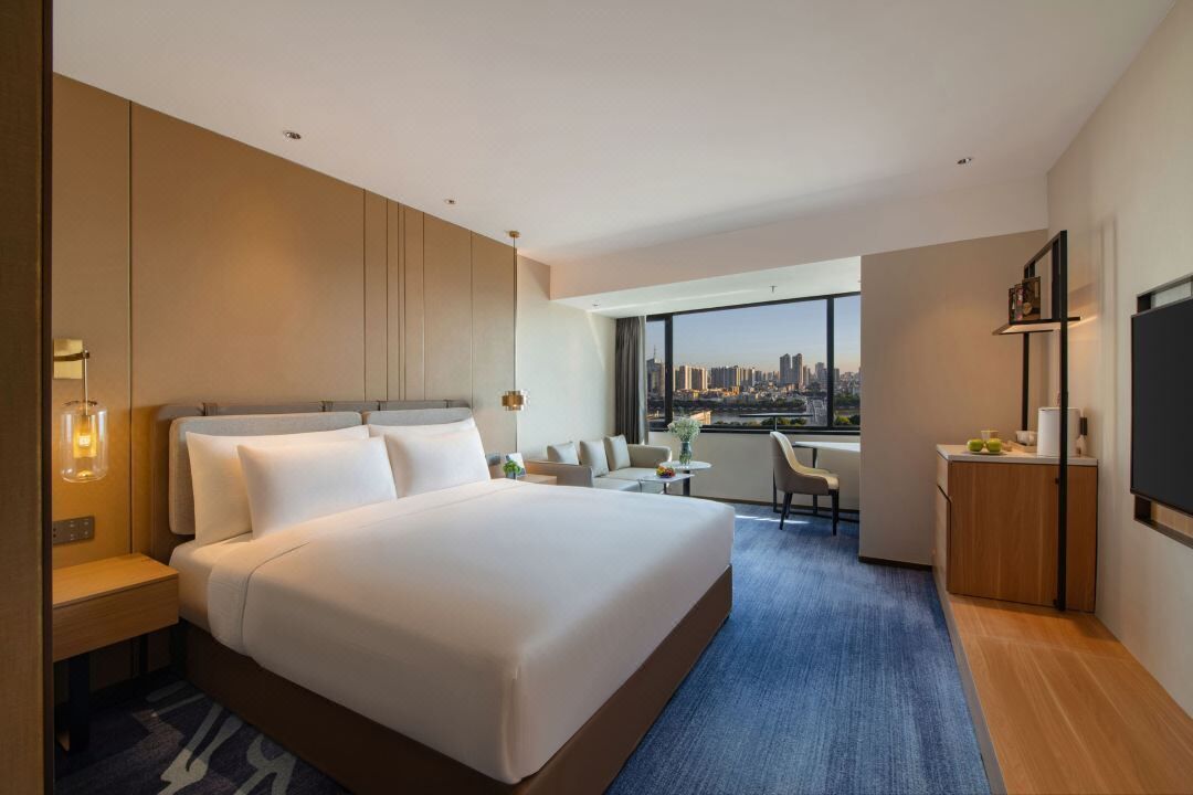 A modern hotel room with a large bed, a desk, a chair, a TV, and a view of a city skyline.