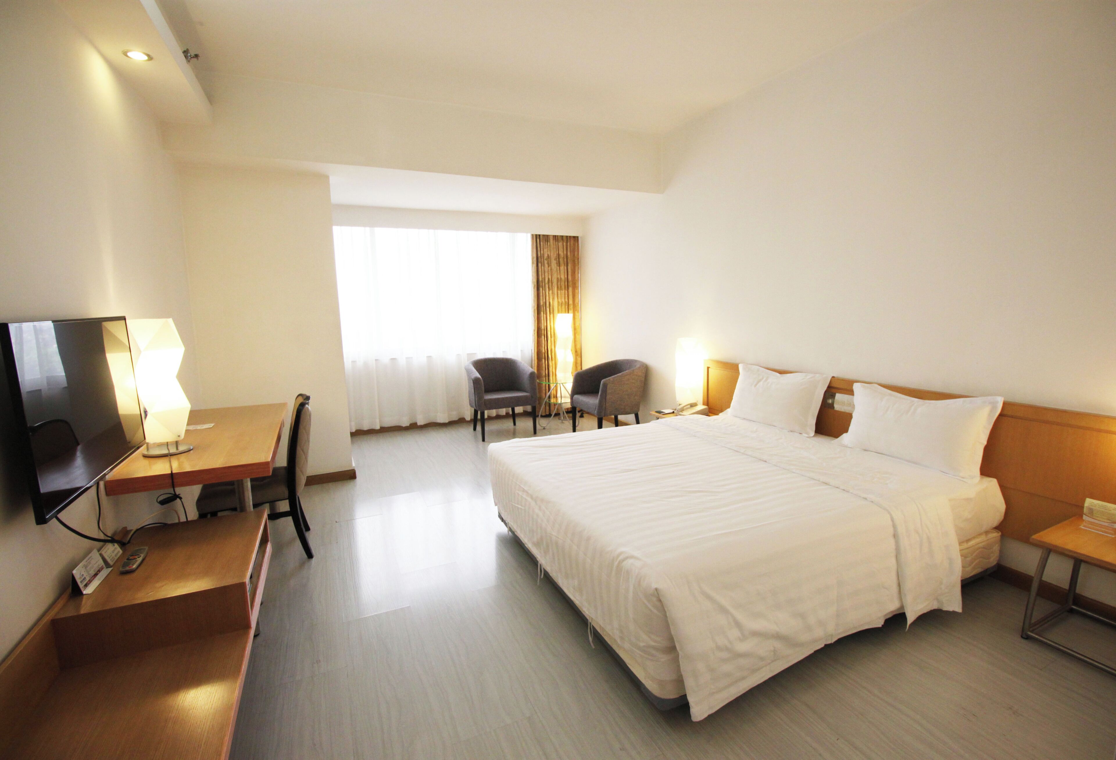 A hotel room with a large bed, a desk with a TV, two chairs, and a window with curtains.