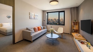 A modern hotel room with a grey sofa, a white armchair, a coffee table, a bookshelf, and a large window with a city view. - Guangzhou Hotel - Canton Fair Free Shuttle Bus (Guangzhou)