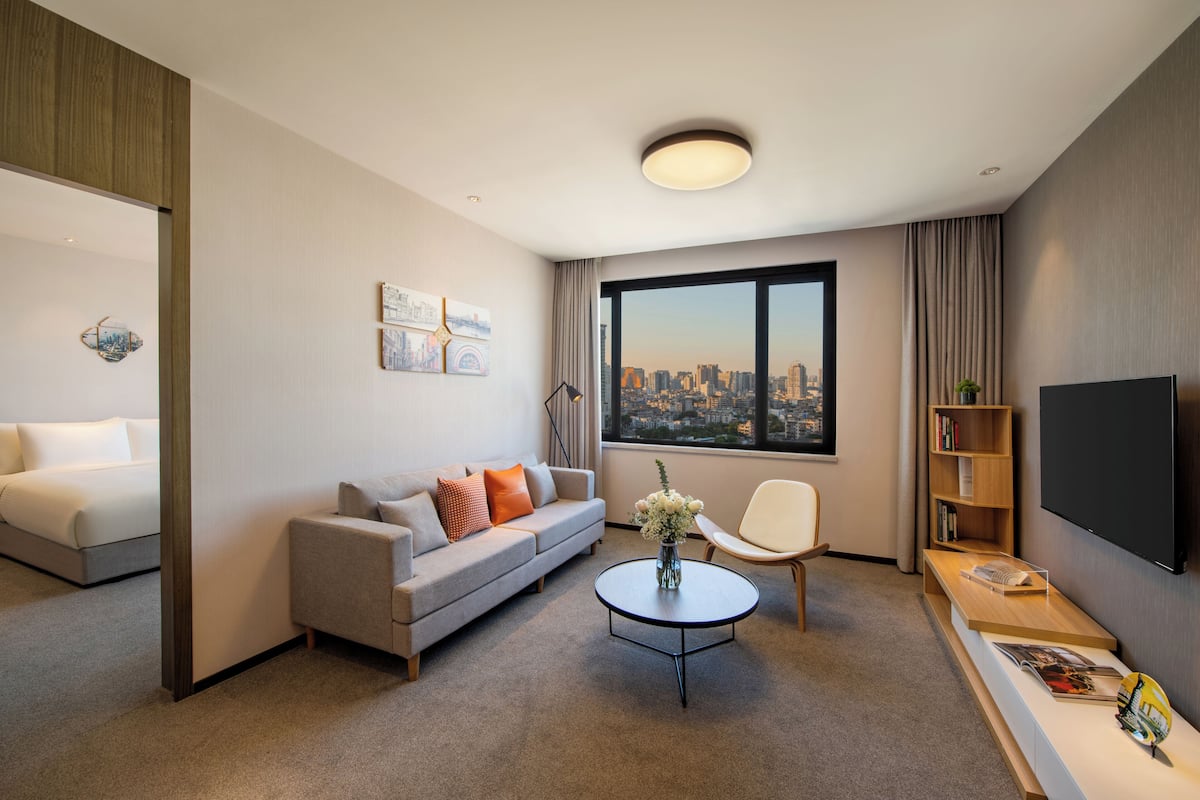 Una habitación de hotel moderna con un sofá gris, un sillón blanco, una mesa de centro, una estantería y un amplio ventanal con vistas a la ciudad.