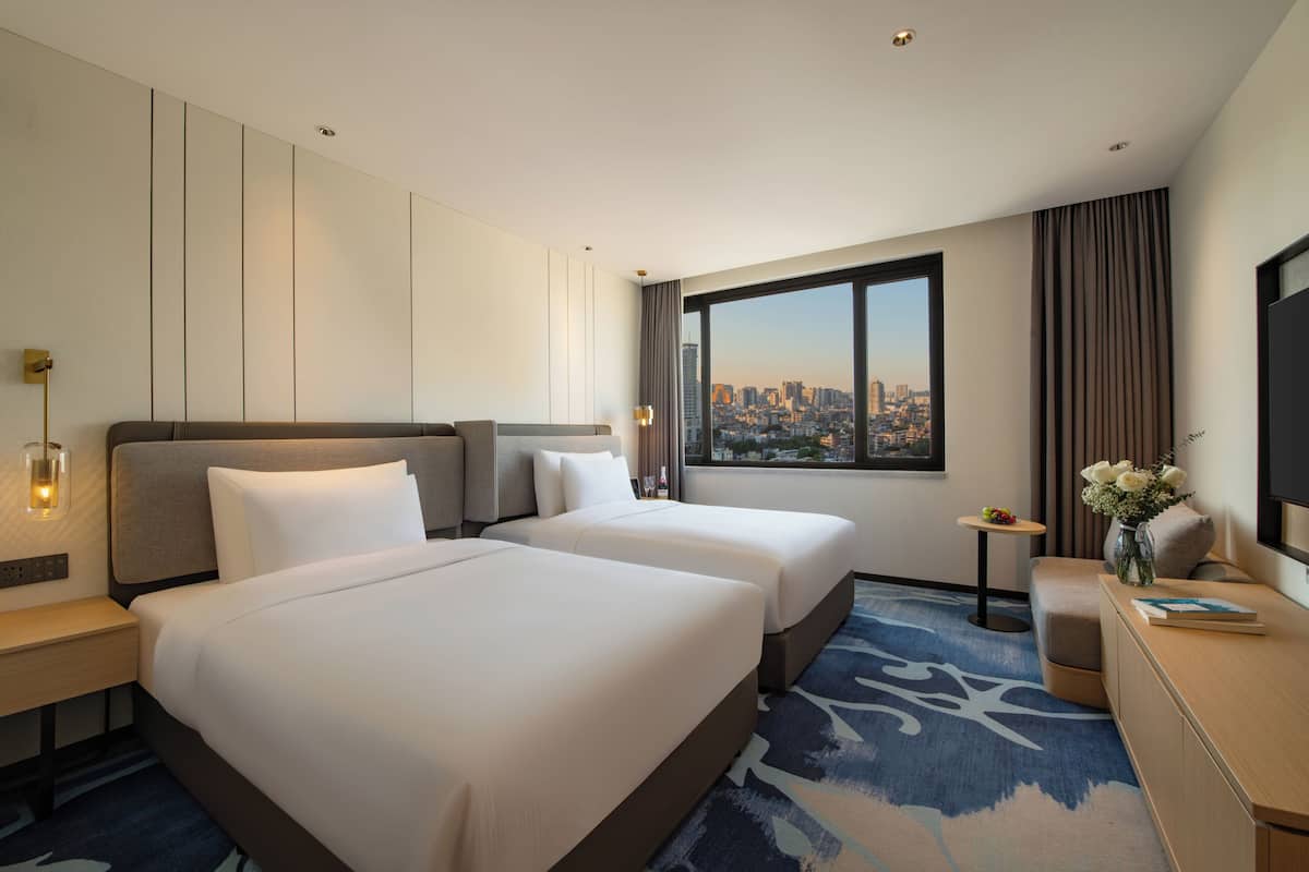 Habitación de hotel con dos camas, un escritorio, una silla, una mesita con flores y un ventanal con vistas a la ciudad.