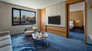 A hotel room with a large window offering a city view, a flat-screen TV mounted on the wall, a small round table with a vase of flowers, a magazine, and a bottle of wine. - Guangzhou Hotel - Canton Fair Free Shuttle Bus (Guangzhou)