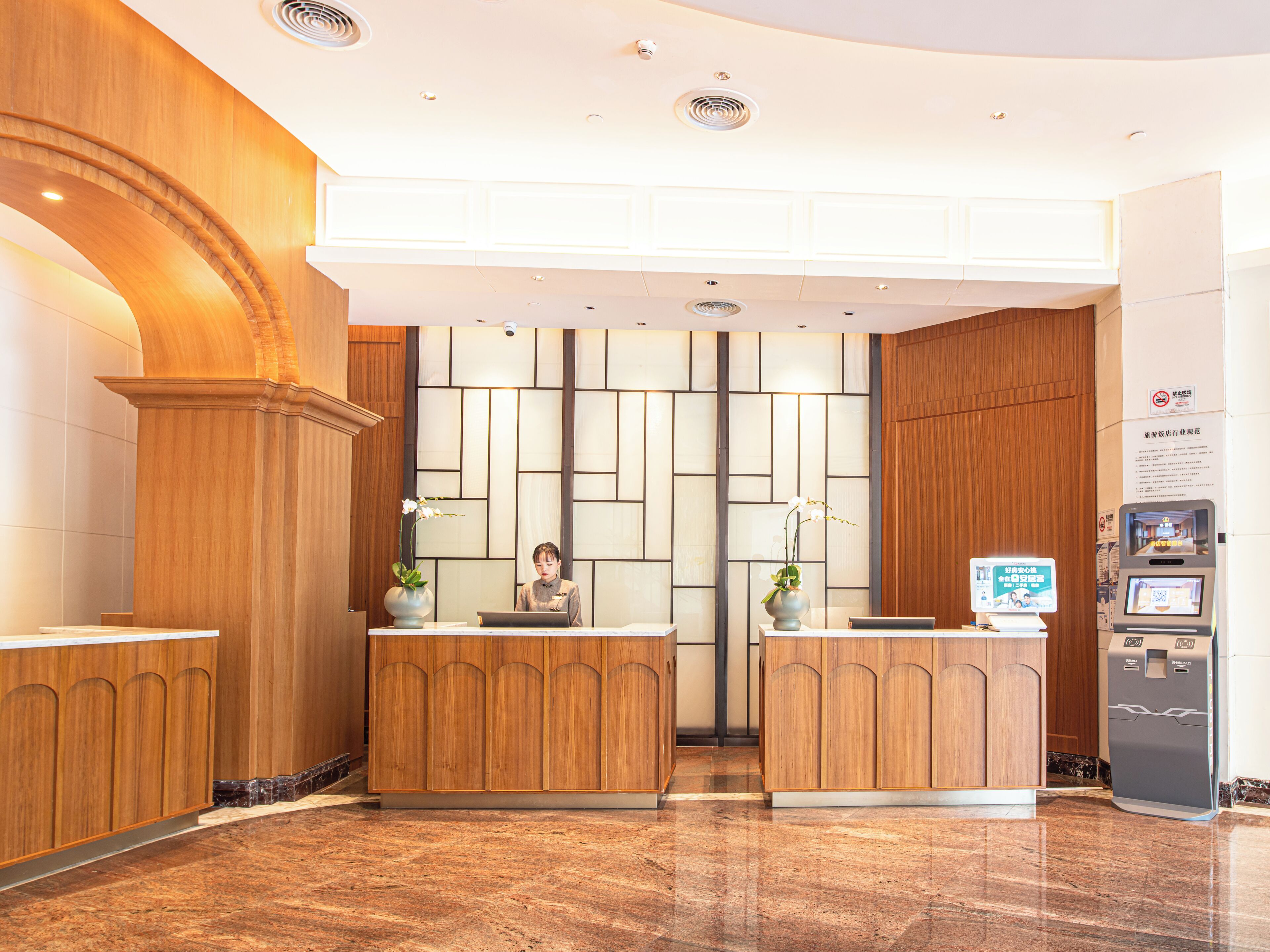 A reception area with wooden reception desks, a receptionist, a digital kiosk, and a tiled floor.