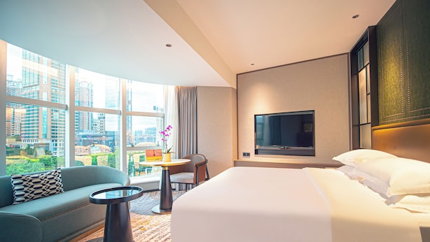 Una habitación de hotel moderna con una cama grande, un sofá, una mesita y un televisor montado en la pared.