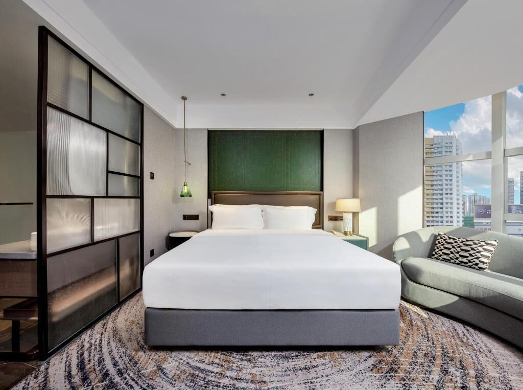 Quarto de hotel moderno com uma cama grande, um sofá, vista da cidade e uma parede de destaque verde.