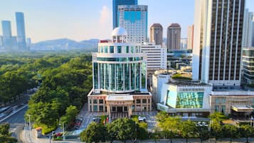 A modern hotel with a glass facade, surrounded by lush greenery and other high-rise buildings.