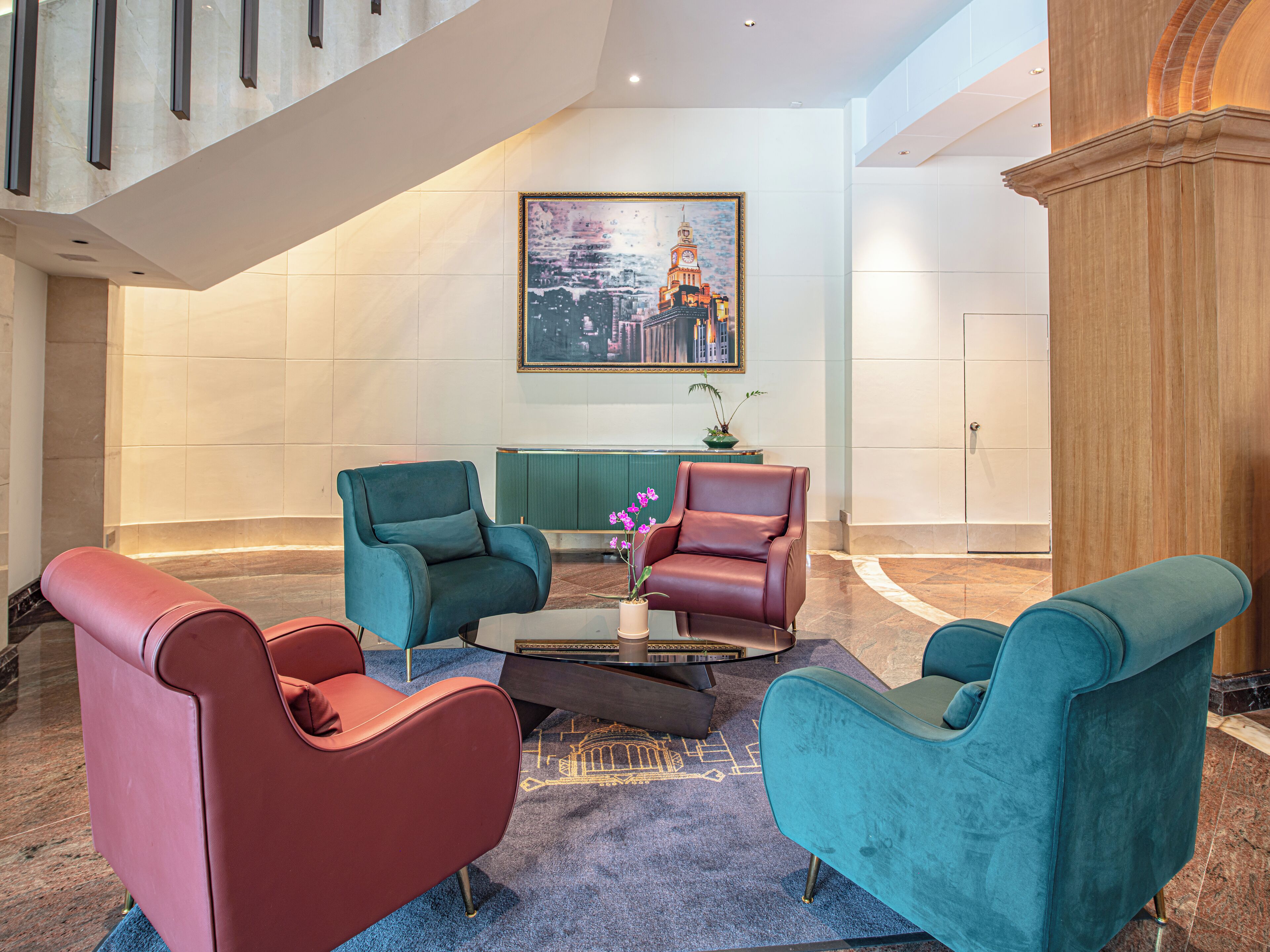 A lobby with a modern design, featuring a seating area with teal and red chairs, a coffee table, and a large painting of a cityscape on the wall.