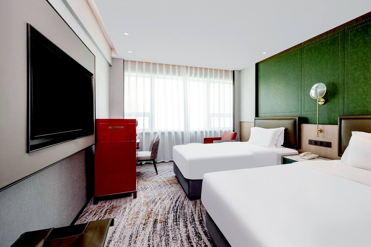 Habitación de hotel con dos camas, un televisor de pantalla plana, un minibar rojo, un escritorio con lámpara y vistas a la ciudad a través de amplias ventanas.