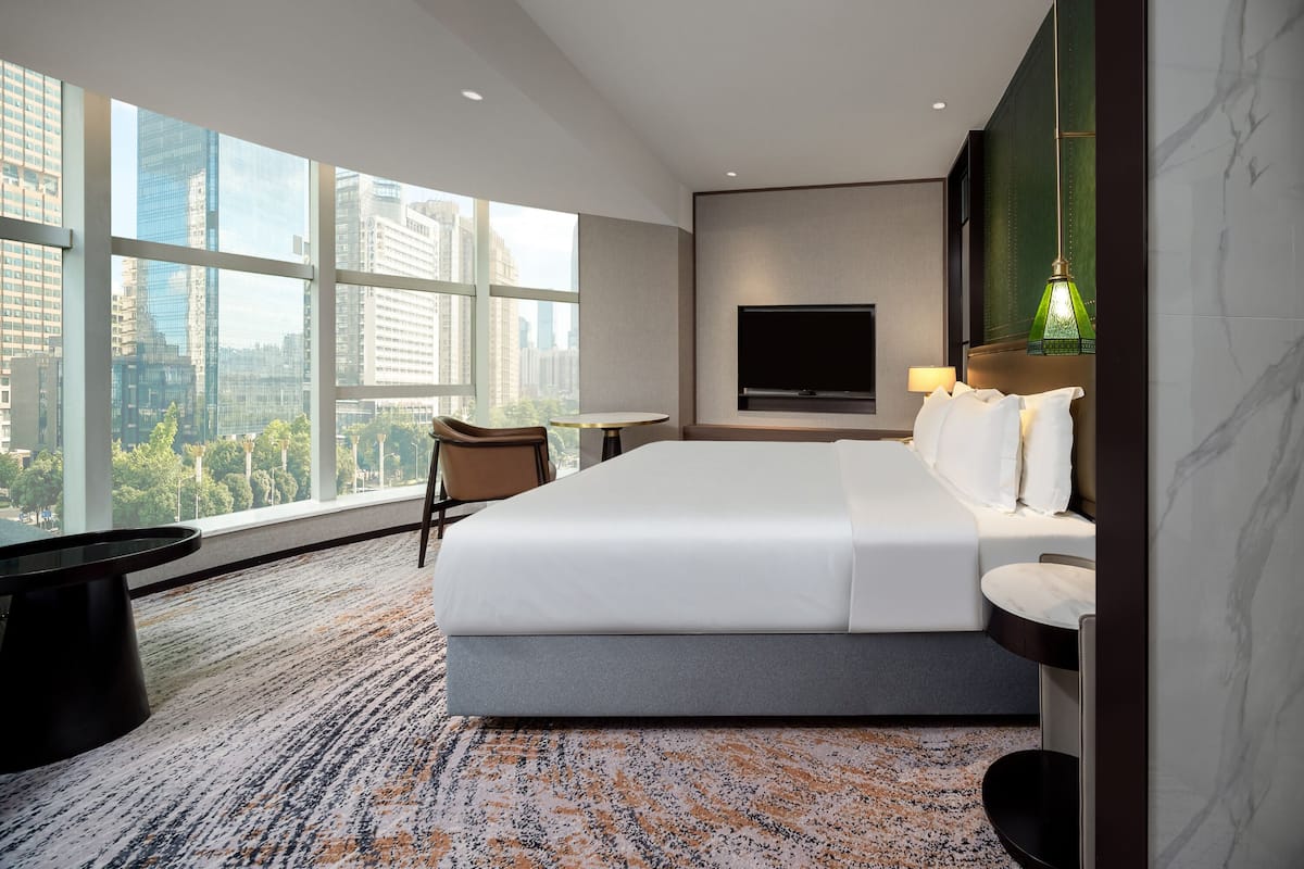 Una habitación de hotel moderna con una cama grande, un televisor montado en la pared, una mesita redonda y vistas de la ciudad a través de amplias ventanas.