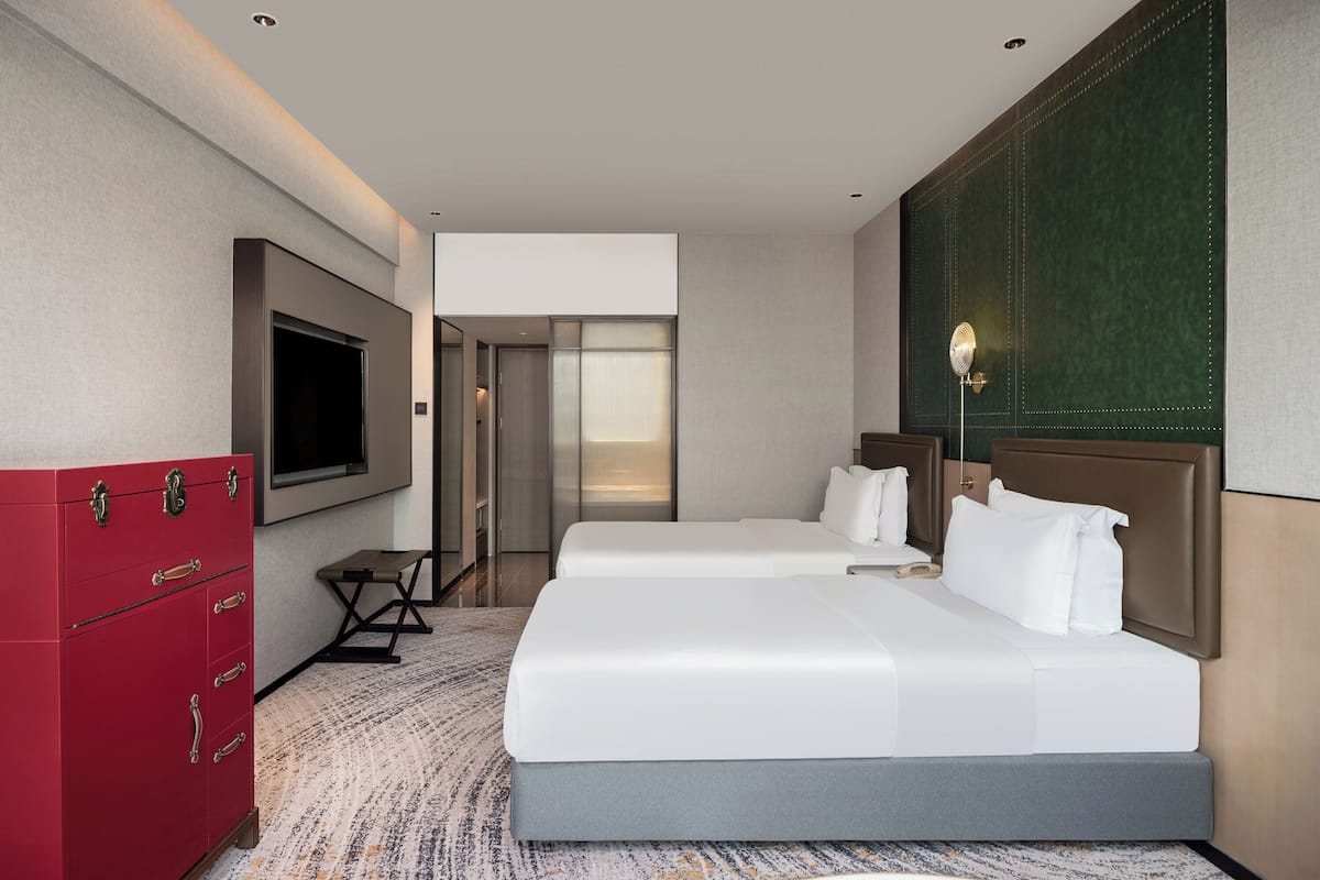 Habitación de hotel con dos camas, televisión, un mueble de almacenamiento rojo y vista al pasillo.