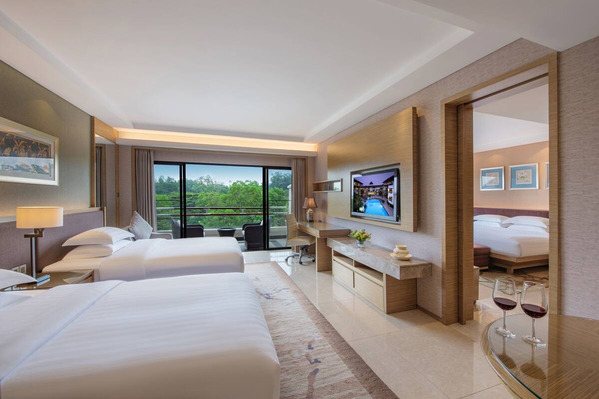 Ein Hotelzimmer mit zwei Betten, einem Schreibtisch, einem Fernseher und einem Balkon mit Blick auf Grünflächen.