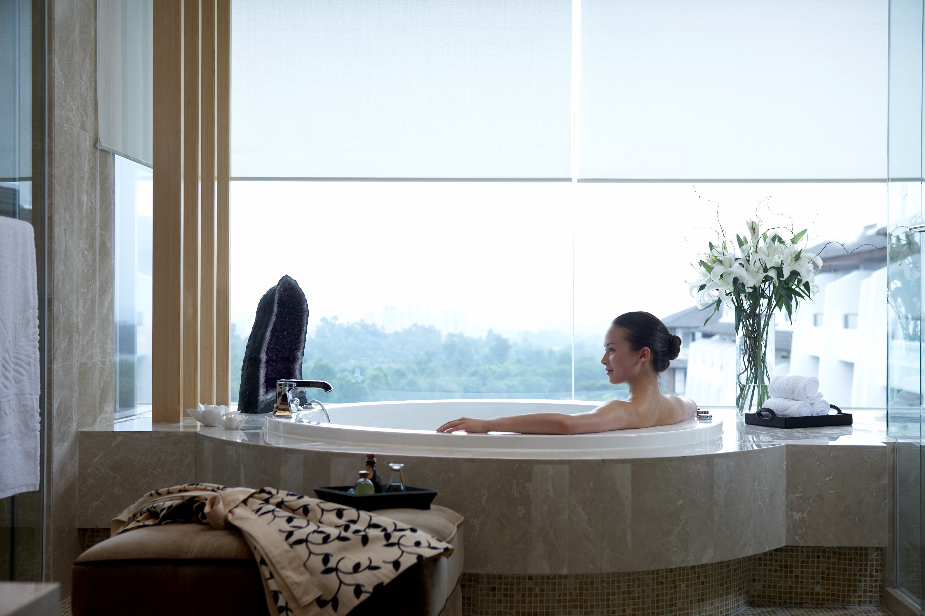 A woman relaxing in a bathtub with a view of a cityscape through a large window.
