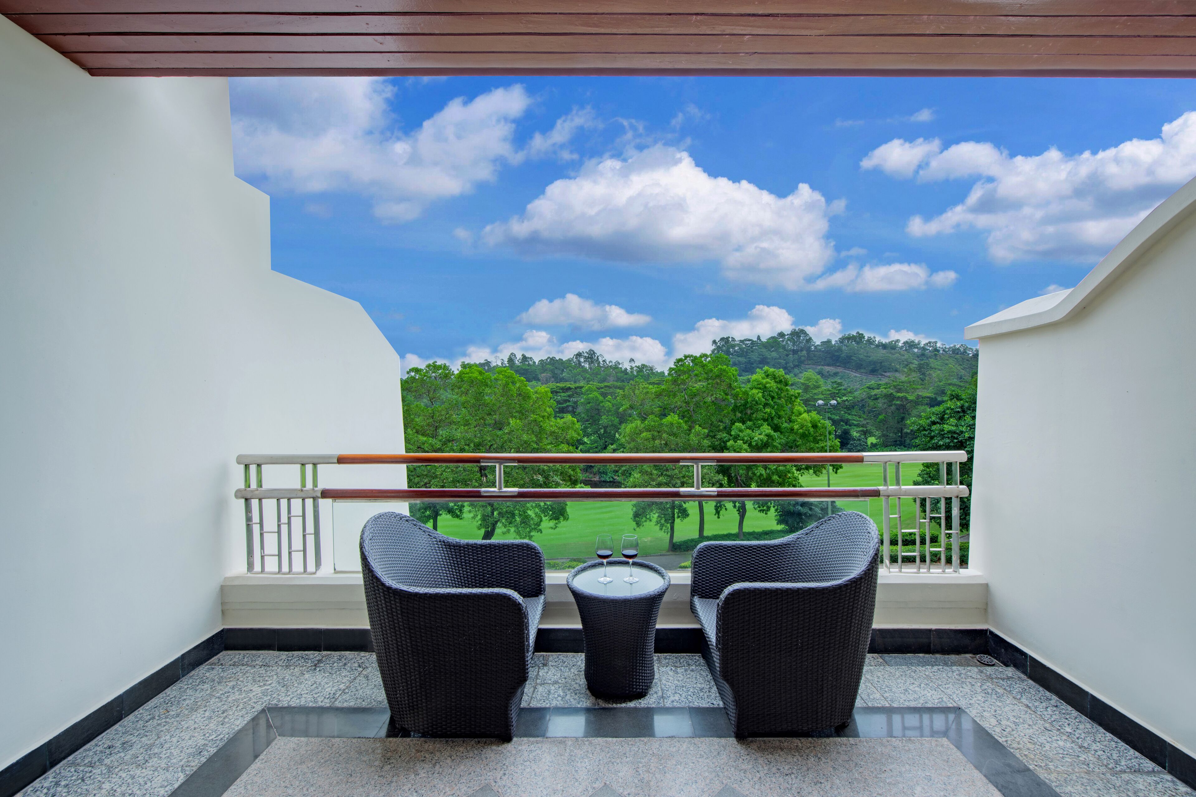 Un balcon avec deux fauteuils en osier et une petite table, d’où l’on peut admirer un paysage verdoyant et un ciel dégagé.