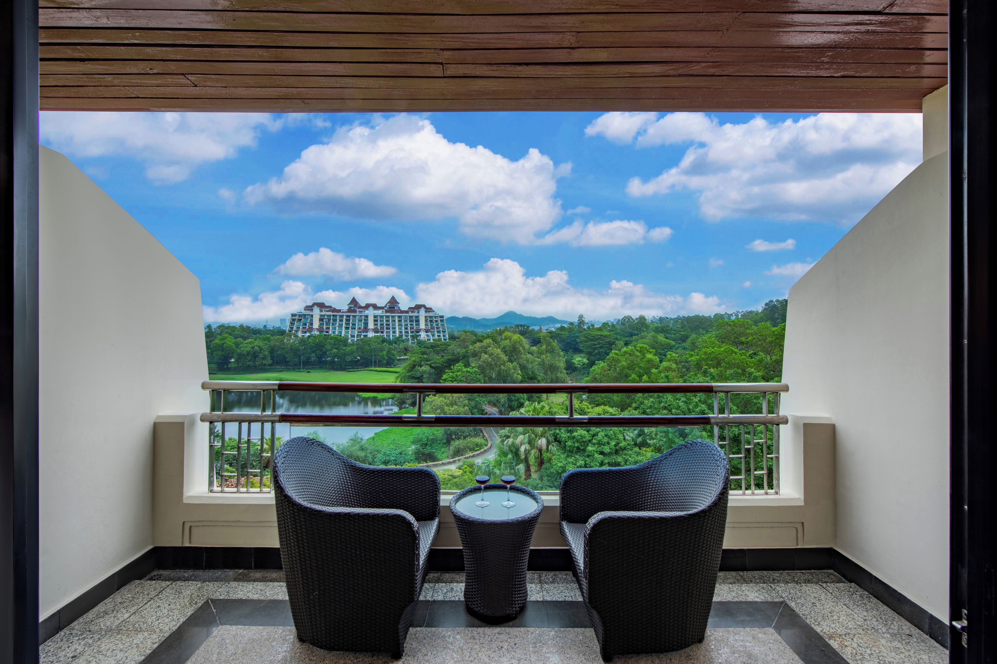 Un balcon agrémenté de fauteuils en osier et d’une petite table, d’où l’on peut admirer un paysage luxuriant et un bâtiment situé au loin.
