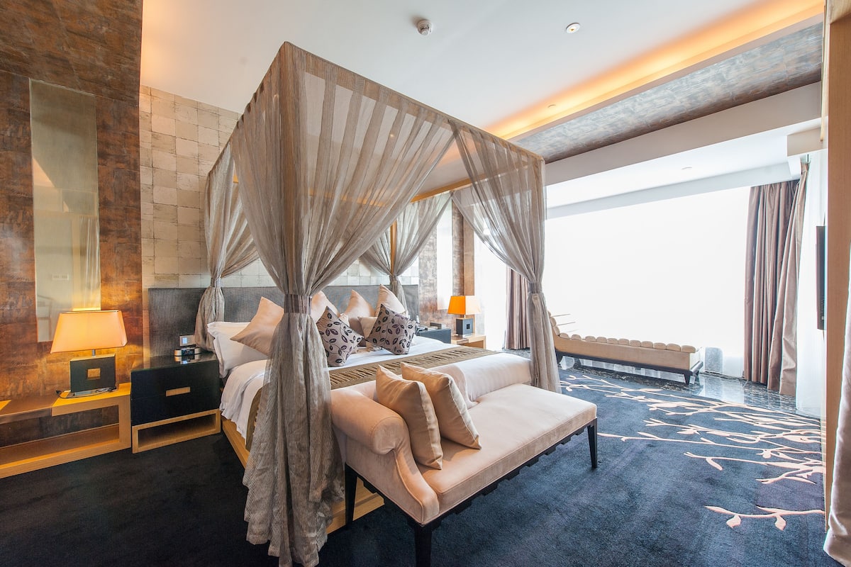Ein geräumiges Schlafzimmer mit Baldachinbett, Nachttisch mit Lampe, einer Bank und Blick auf die Stadt.